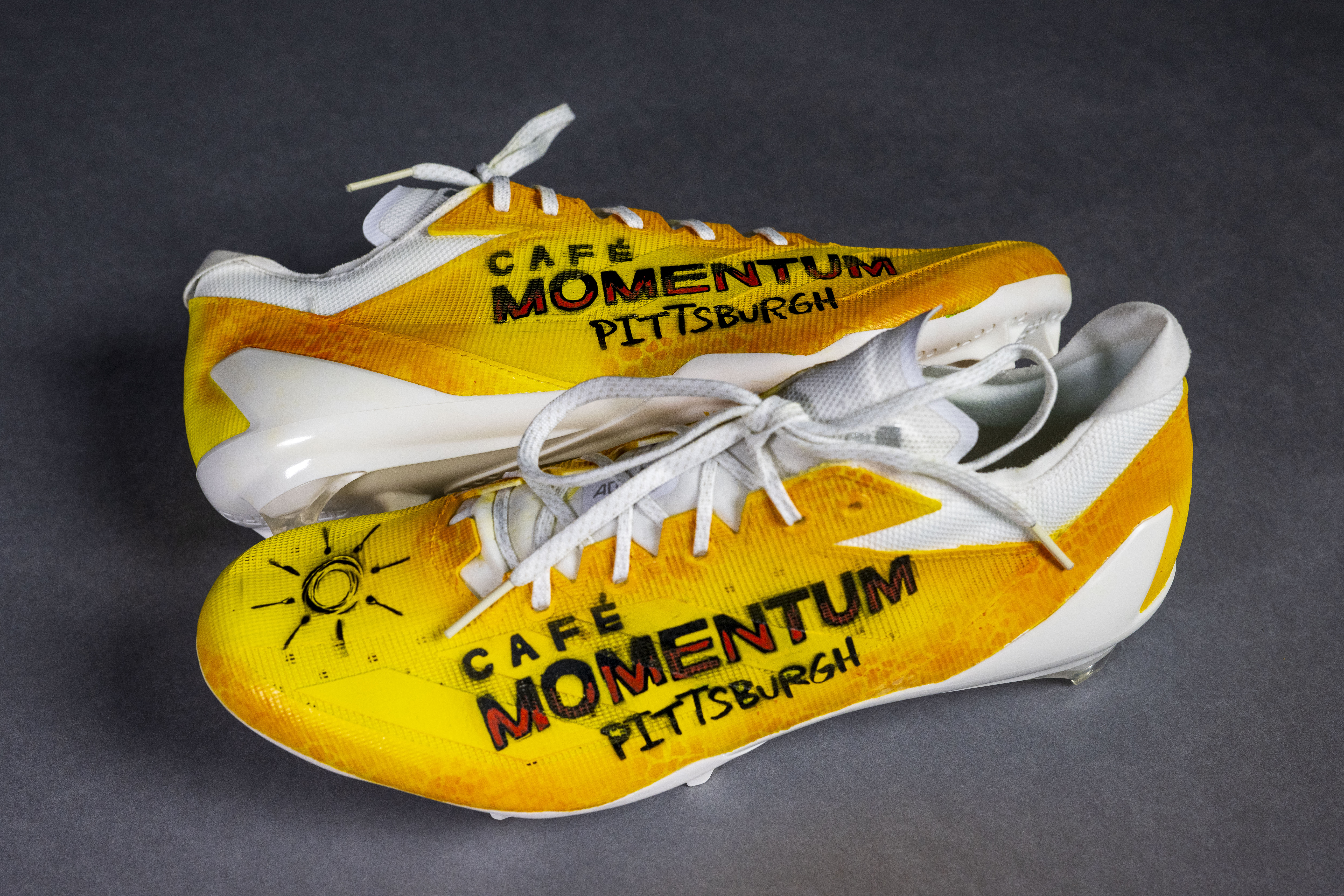 Yellow athletic shoes with "Café Momentum Pittsburgh" printed on them, featuring a sun doodle on the toe area.