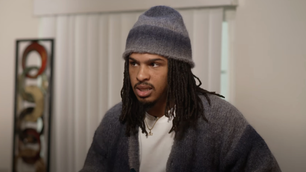 Keith Lee with long hair and a beard wearing a gray beanie and sweater, standing indoors with a neutral expression.