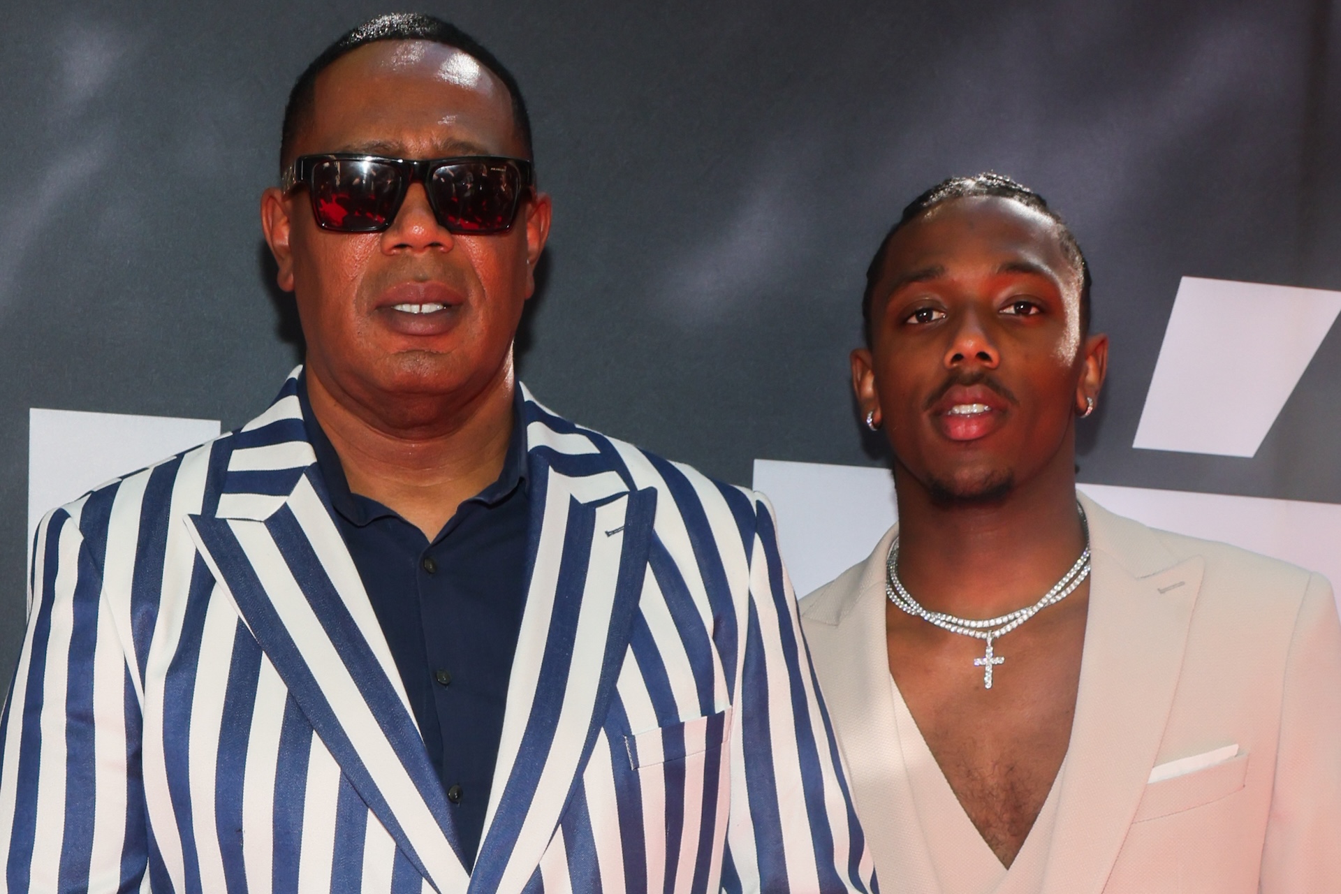 Master P and son Hercy Miller posing together; one wearing a blue and white striped suit and sunglasses, the other in a light-colored suit with a necklace.