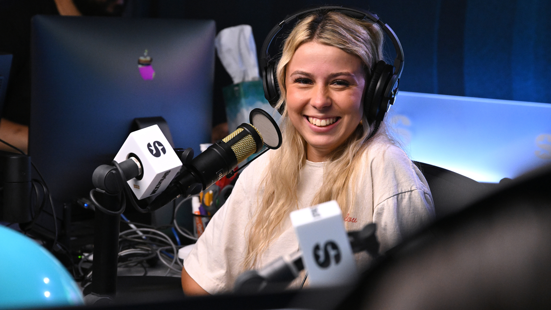 A woman with long blonde hair, wearing headphones, smiles while sitting at a radio studio microphone.
