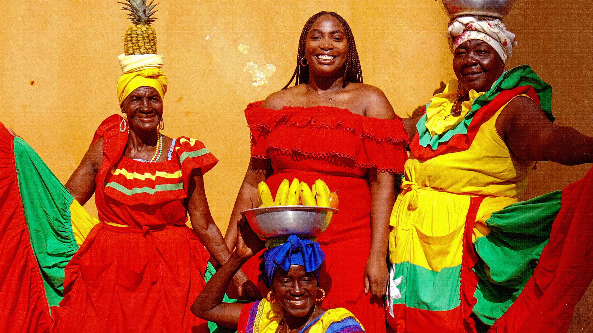 Alexa Moore and three women in vibrant traditional attire, each with fruit on their heads, smiling in front of an orange wall.