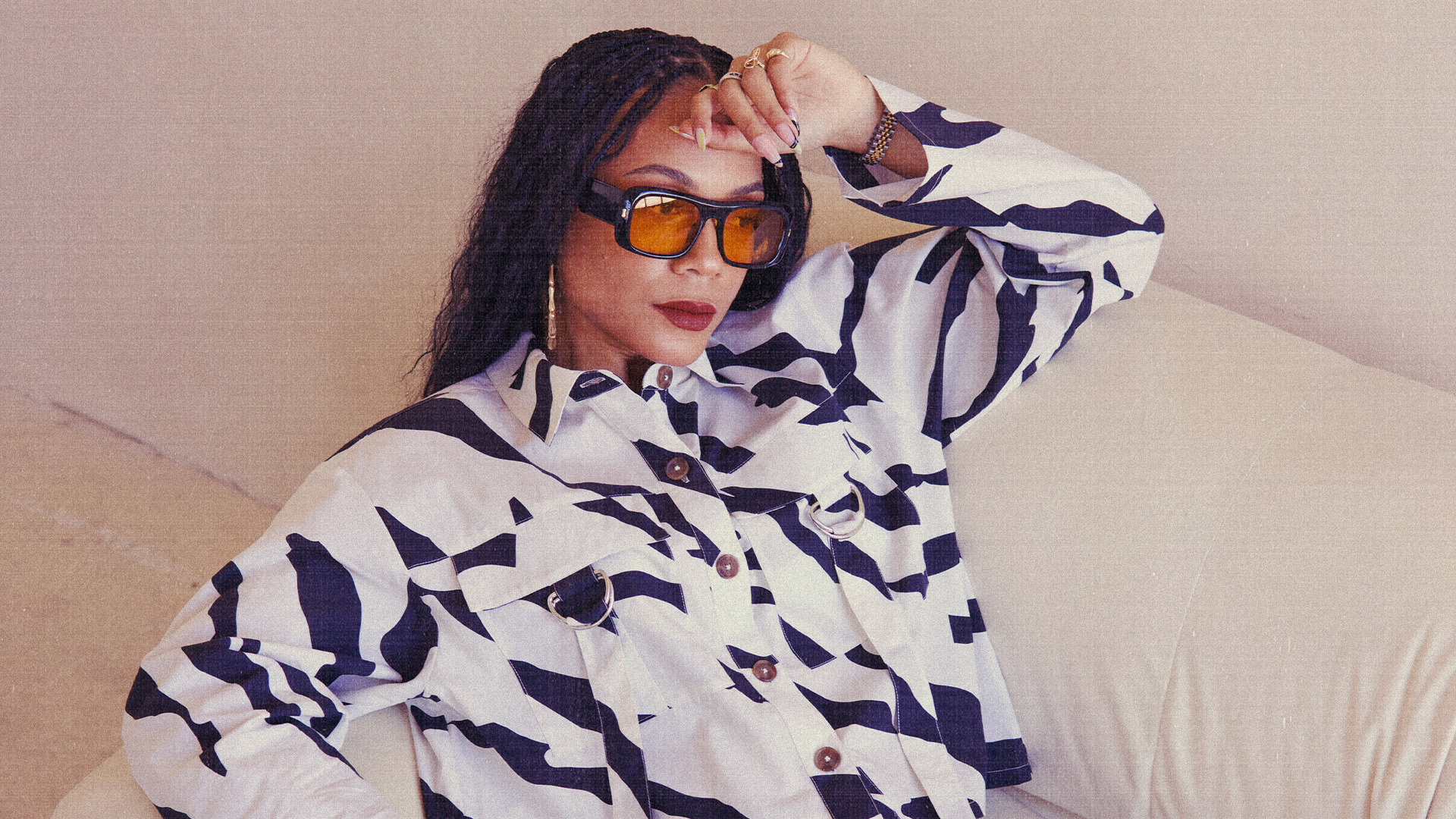Kia Marie in a black and white patterned jacket, wearing sunglasses, sitting on a couch with a relaxed pose.