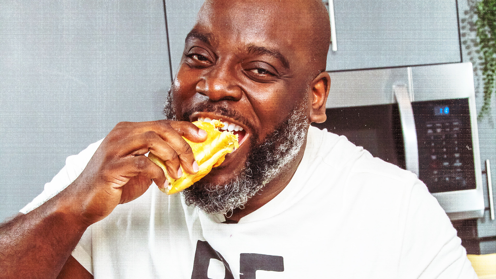 Darius Williams is smiling while eating a sandwich in a kitchen. He is wearing a white t-shirt.