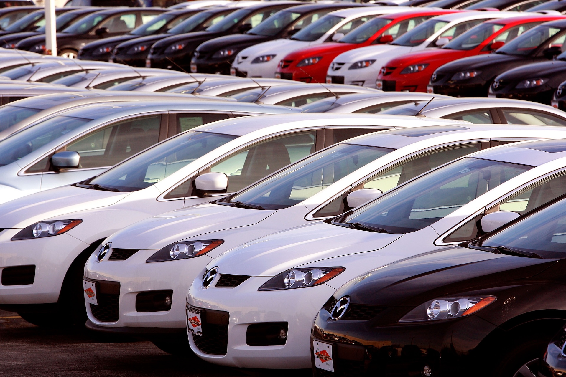 Rows of parked cars in various colors, including white, black, and red, at a dealership lot.