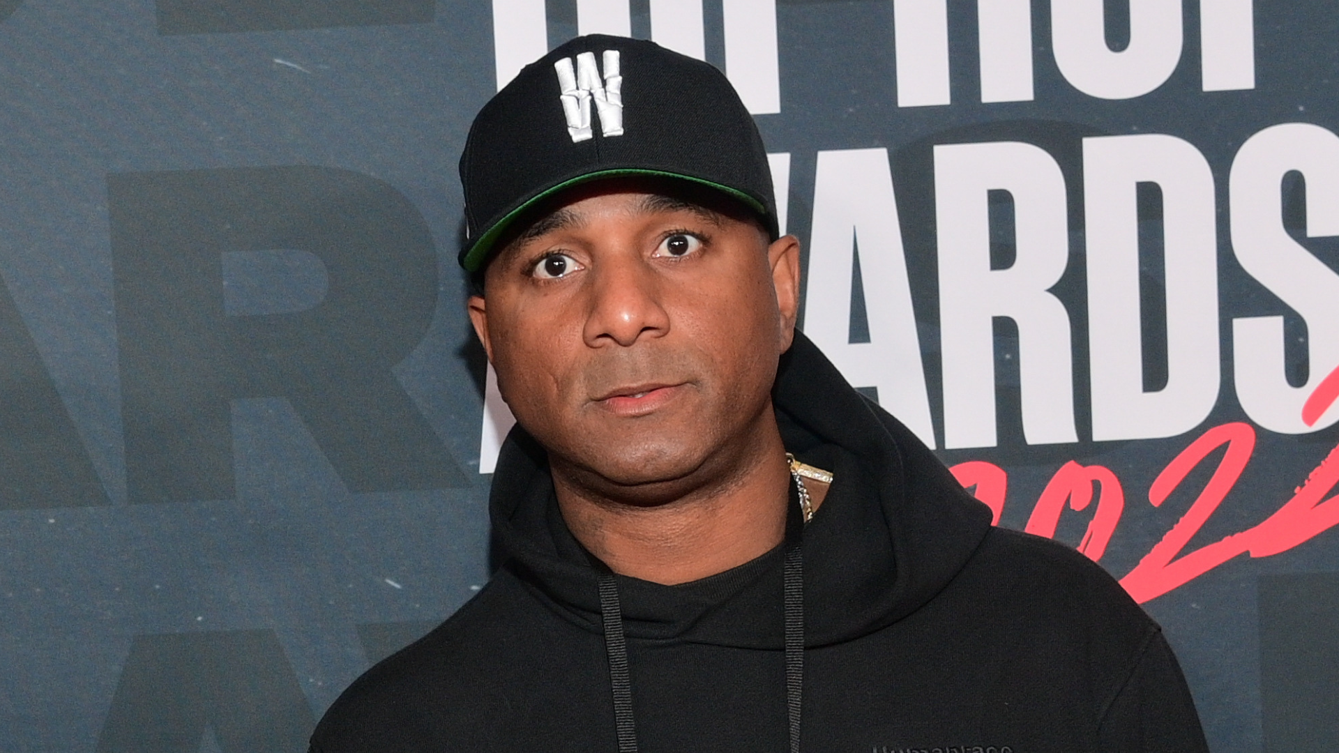 A man wearing a black hoodie and cap stands in front of a backdrop with "Hip Hop Awards" text.