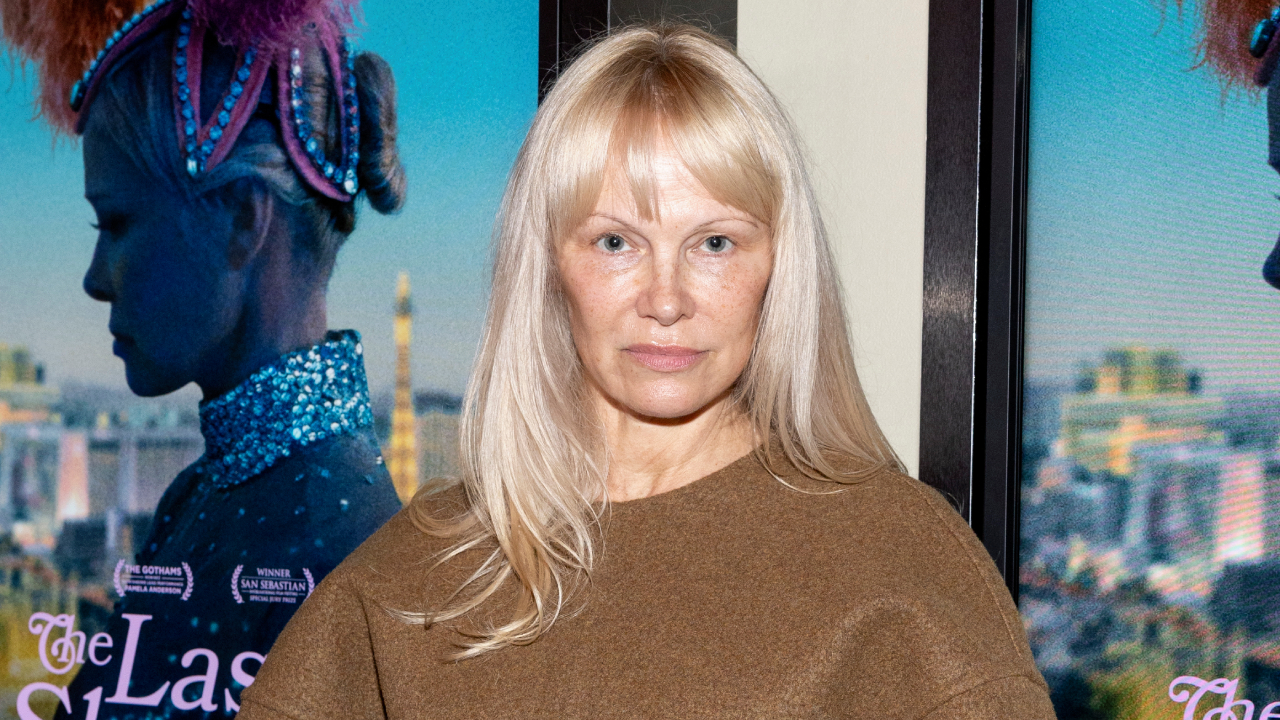 Pamela Anderson with long blonde hair stands in front of a movie poster. The background features a cityscape and a performer.
