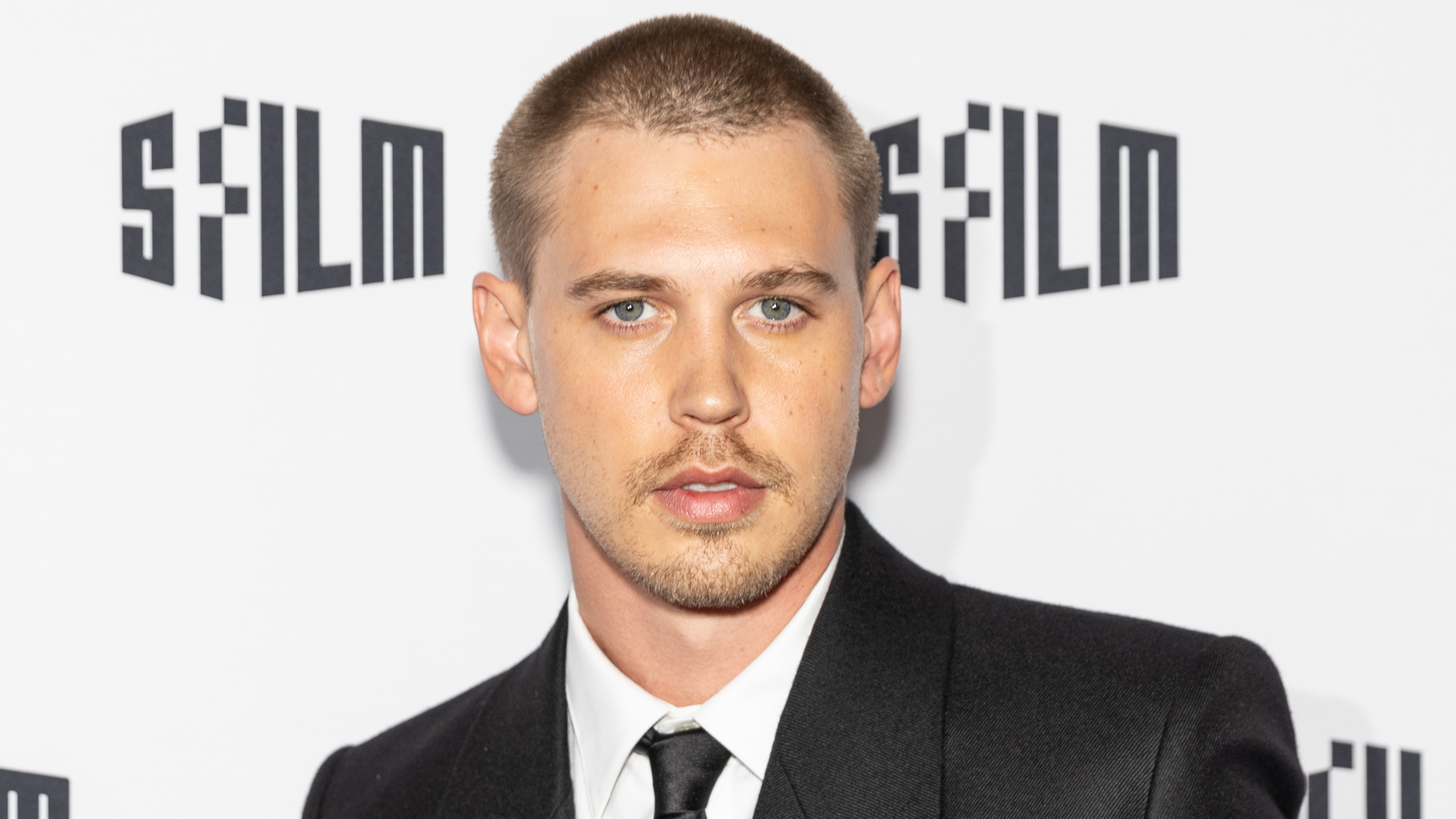 Austin Butler in a black suit and tie at an event, posing against a white background.