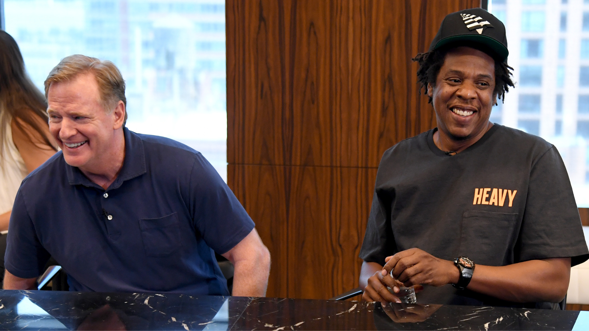 Roger Goodell and Jay-Z sitting at a table, smiling. Jay-Z wears a "Heavy" shirt and cap.