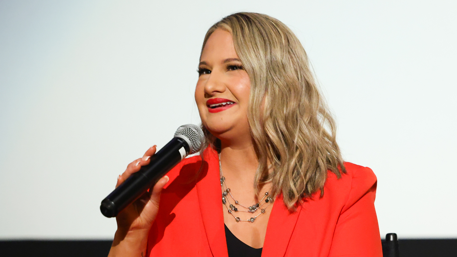 Gypsy Rose Blanchard in a red blazer speaks into a microphone while seated on a stage.