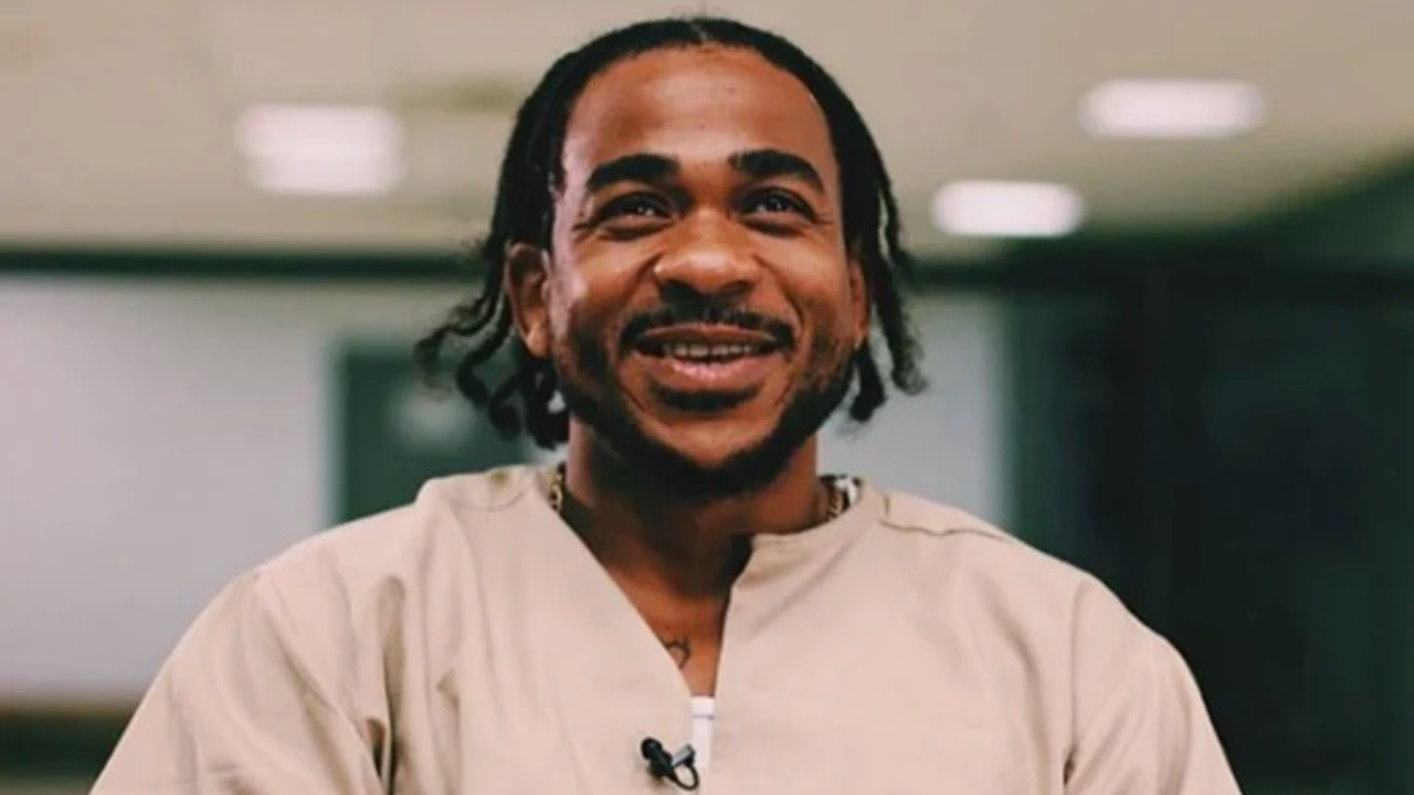Max B with braided hair, wearing a beige shirt, smiles in a well-lit room.
