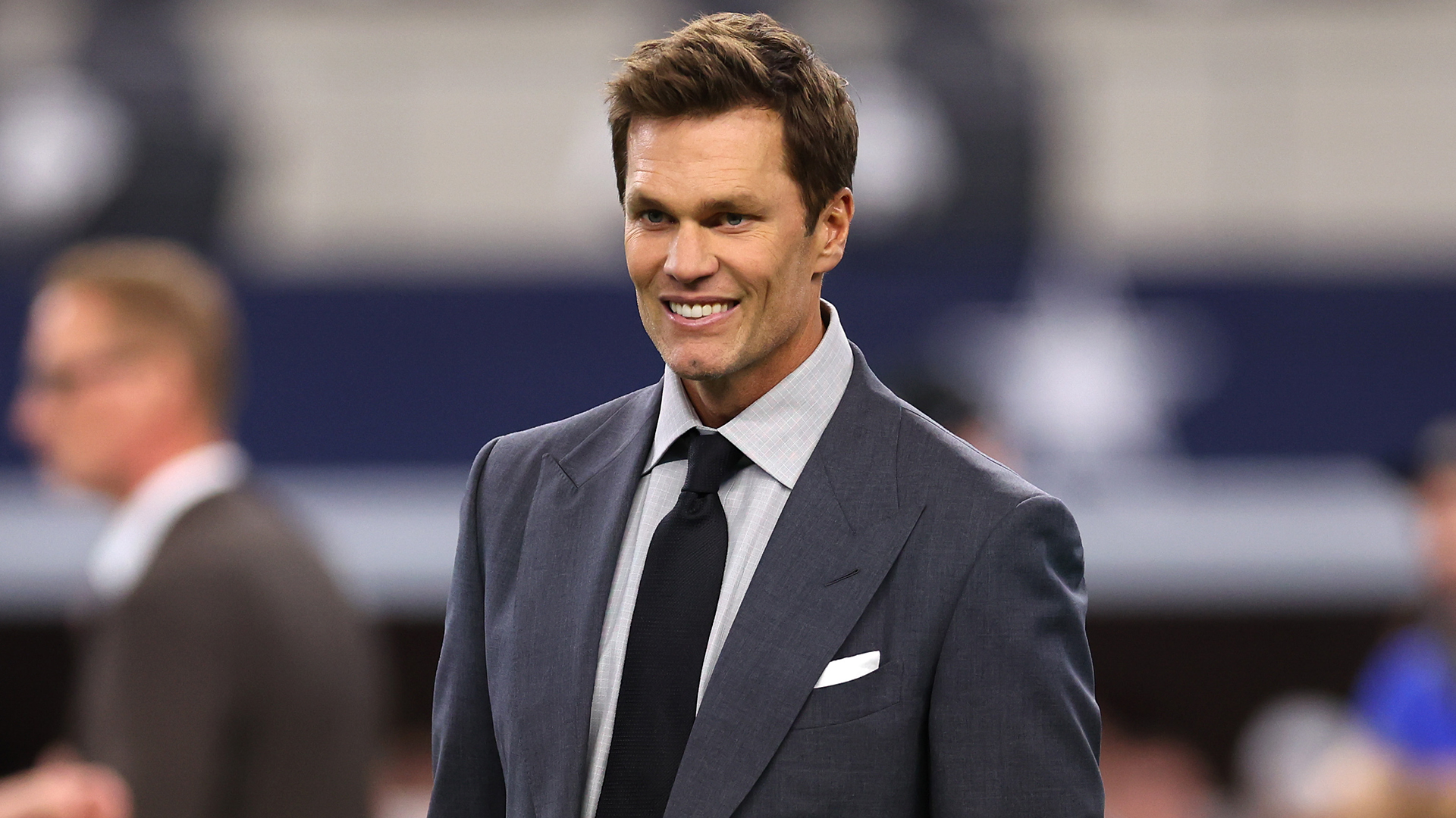 Tom Brady in a suit, smiling at an event. He has short hair and is wearing a dark tie.