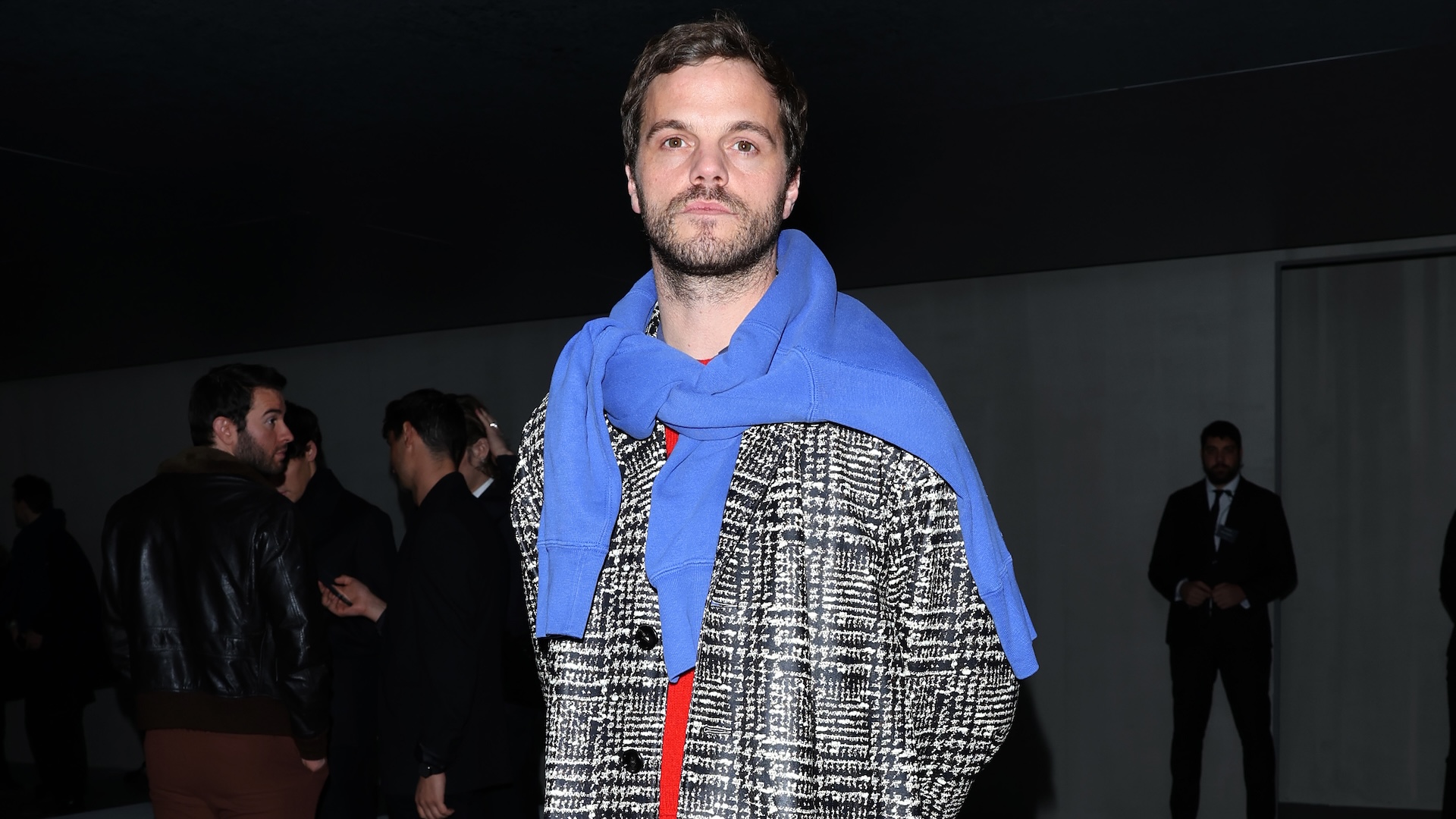 Matthieu Blazy wearing a plaid coat with a blue sweater draped over his shoulders stands in a dimly lit room with people in the background.