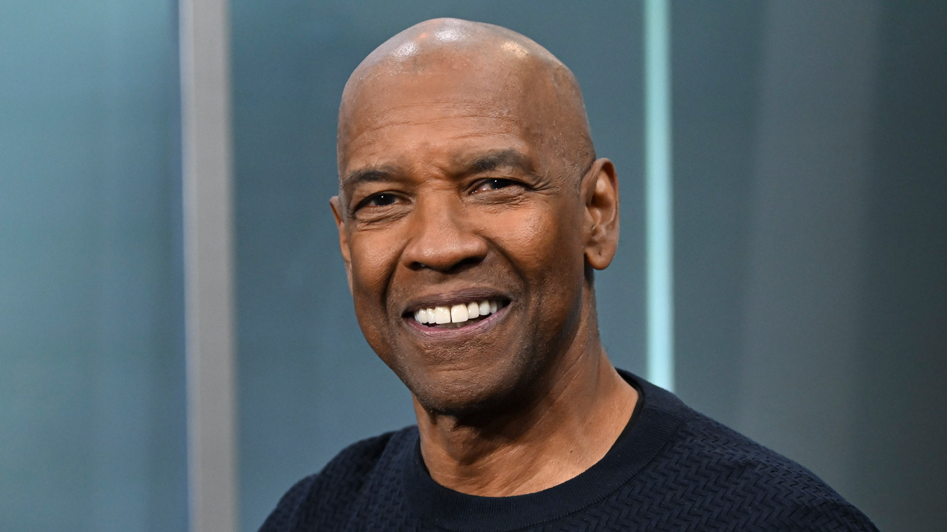 Denzel Washington smiling, wearing a dark sweater, with a neutral background.