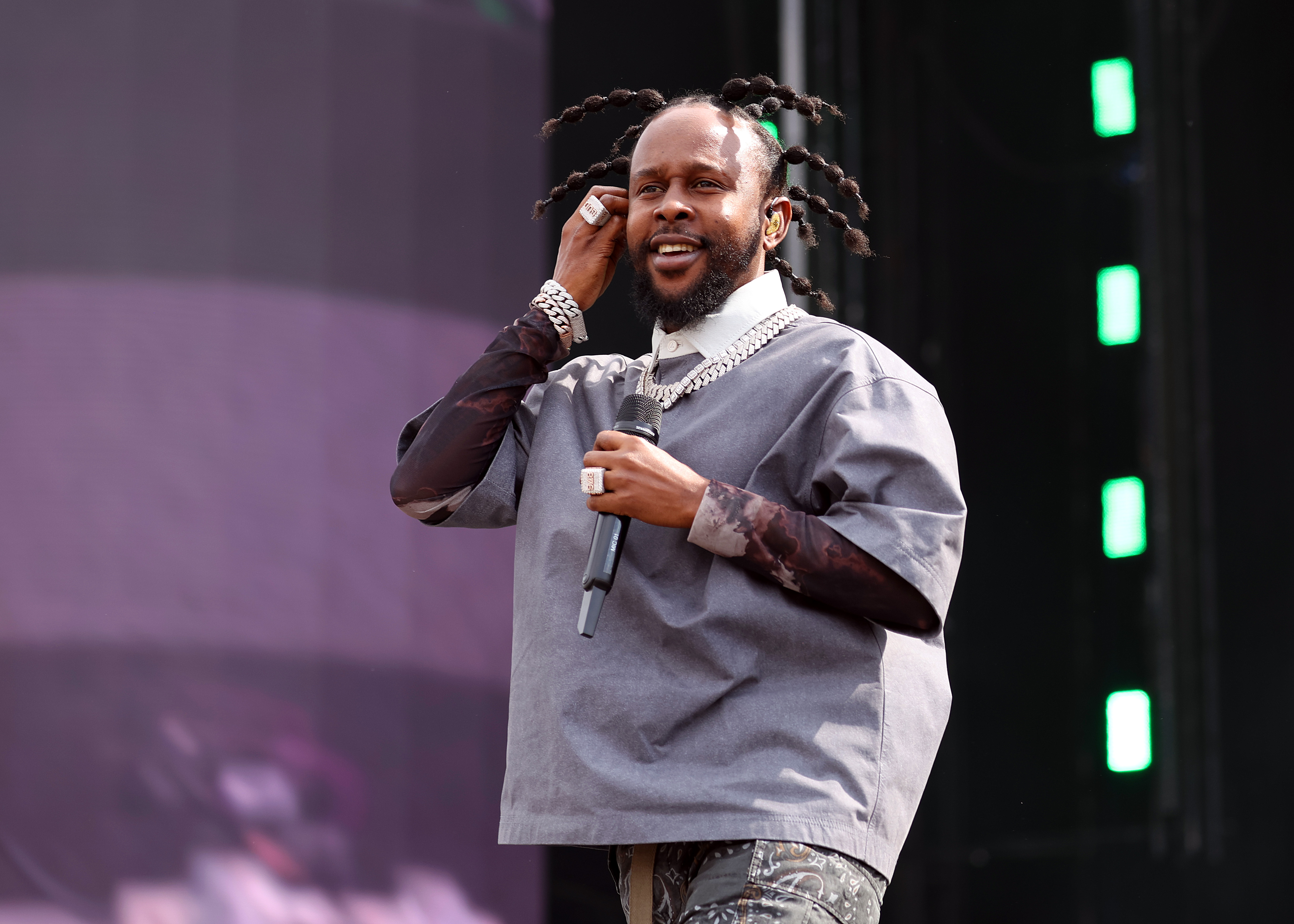 Popcaan braided hair performs on stage, holding a microphone, wearing a gray oversized shirt and jewelry, with a backdrop of lights.