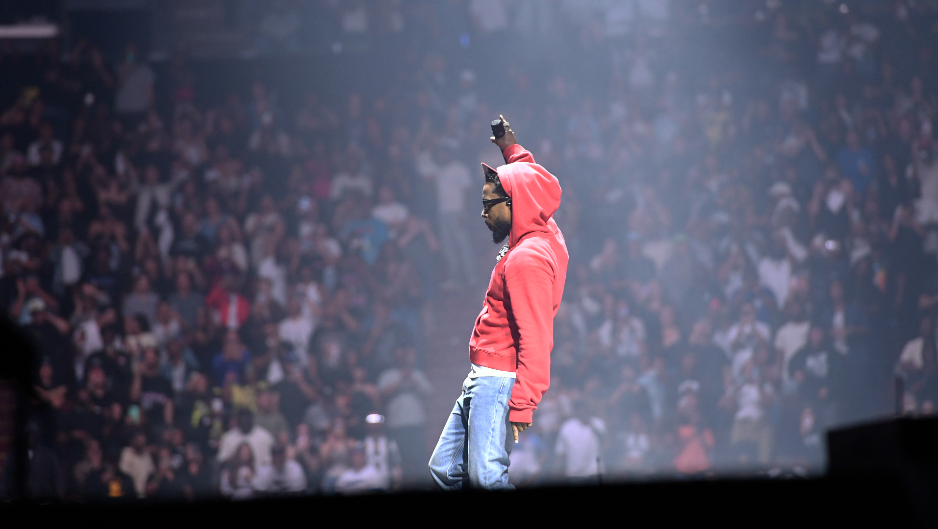 Kendrick Lamar in a red hoodie and jeans on stage, raising one hand. A large crowd is visible in the background.