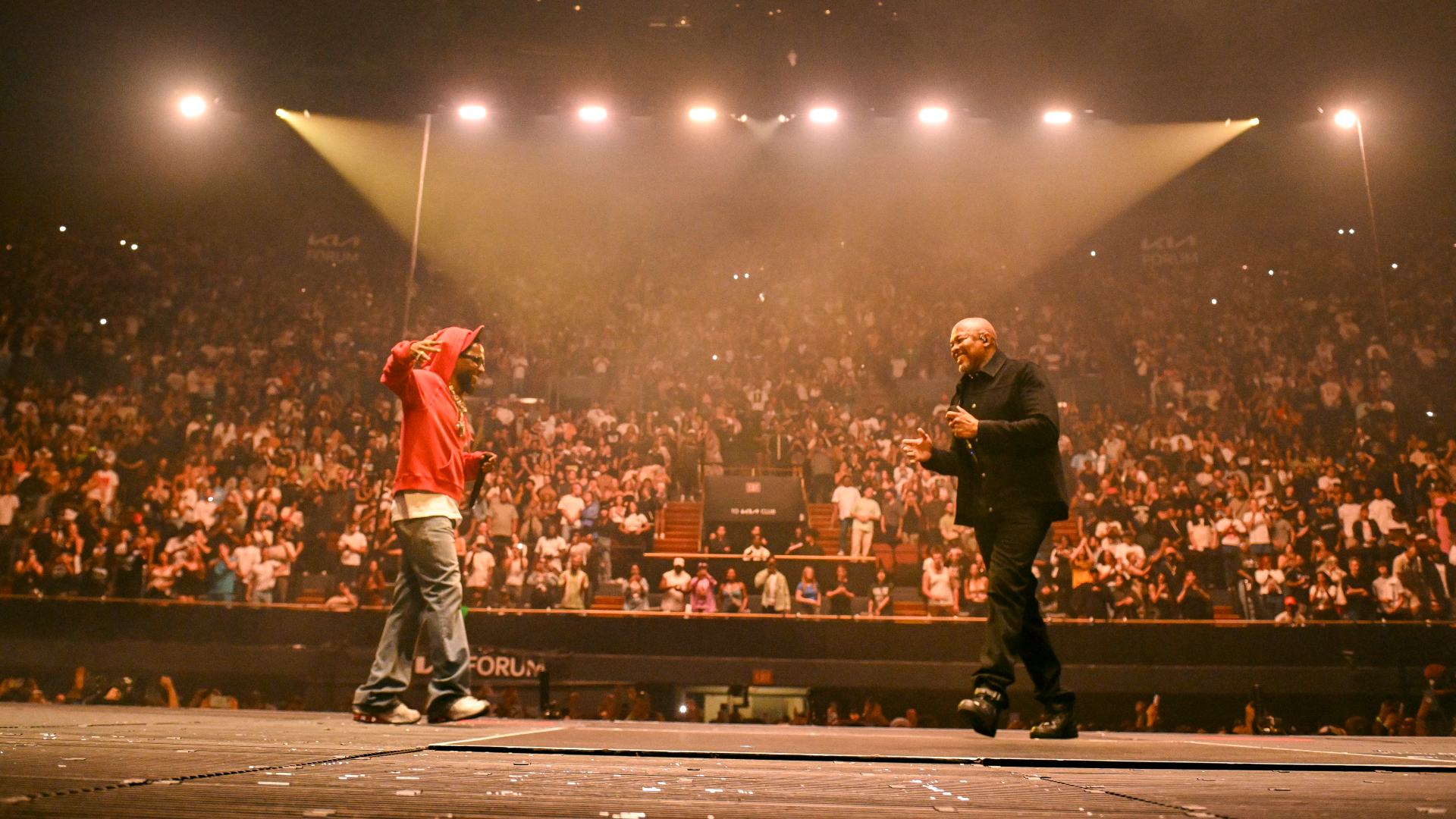Two performers on stage in front of a large audience, with dramatic lighting highlighting the scene.
