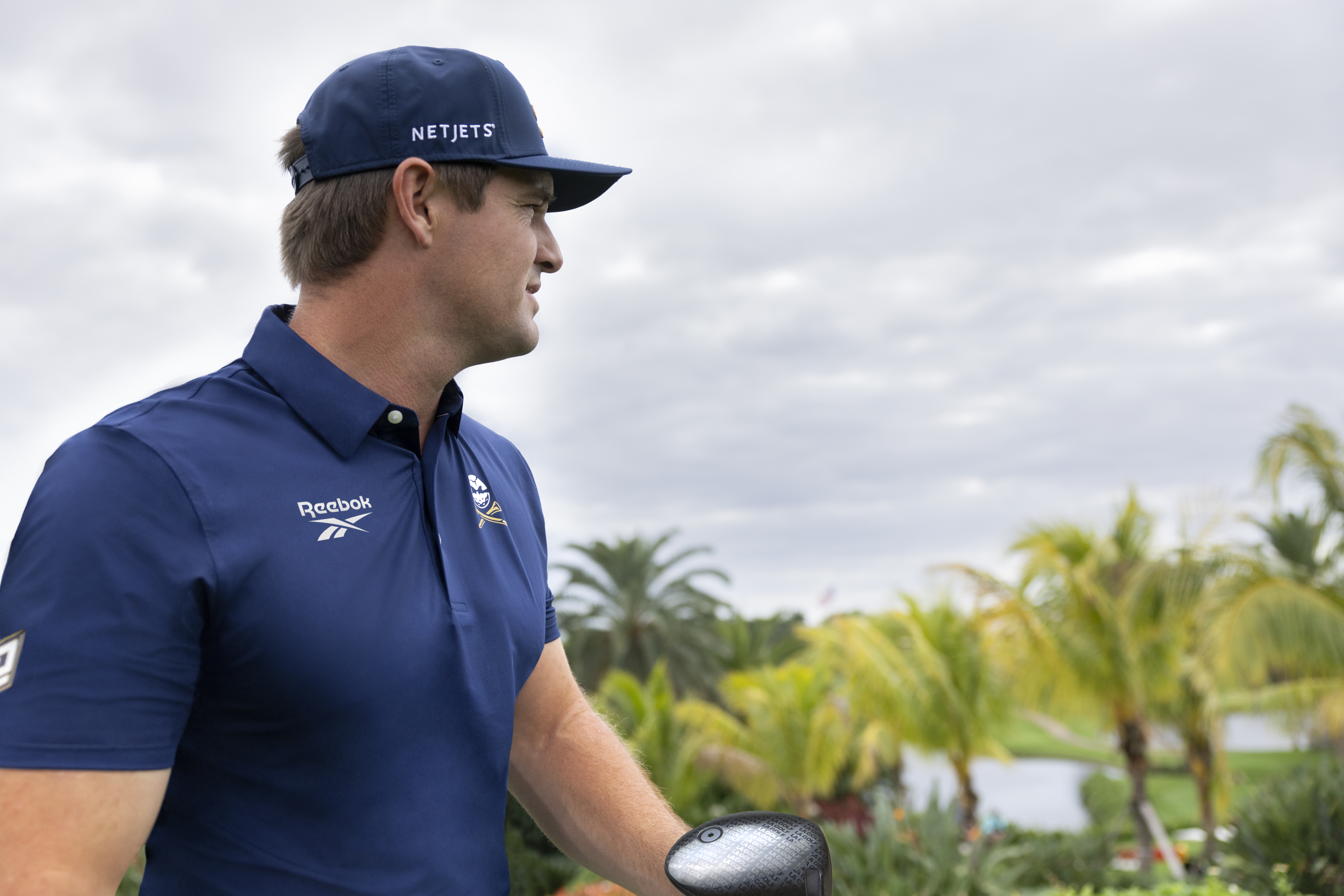 A man in a blue Reebok shirt and cap stands outdoors with palm trees in the background, holding a golf club.