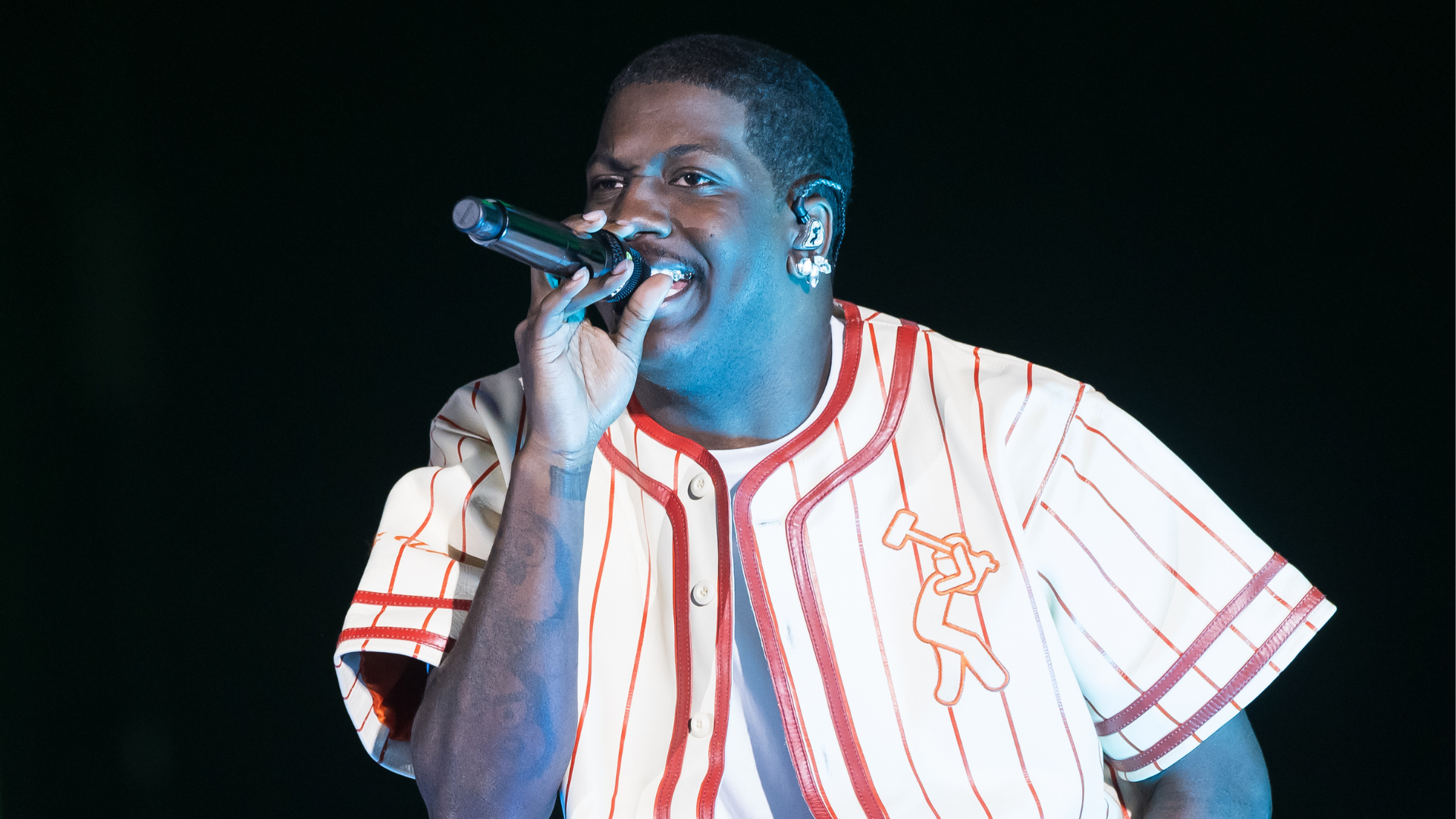 A man performing on stage, wearing a striped baseball-style shirt, holding a microphone, with a focused expression.