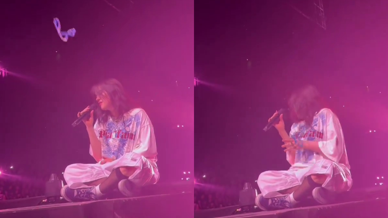 Billie Eilish sitting on stage under pink lighting, holding a microphone, with a bracelet being thrown in the air.