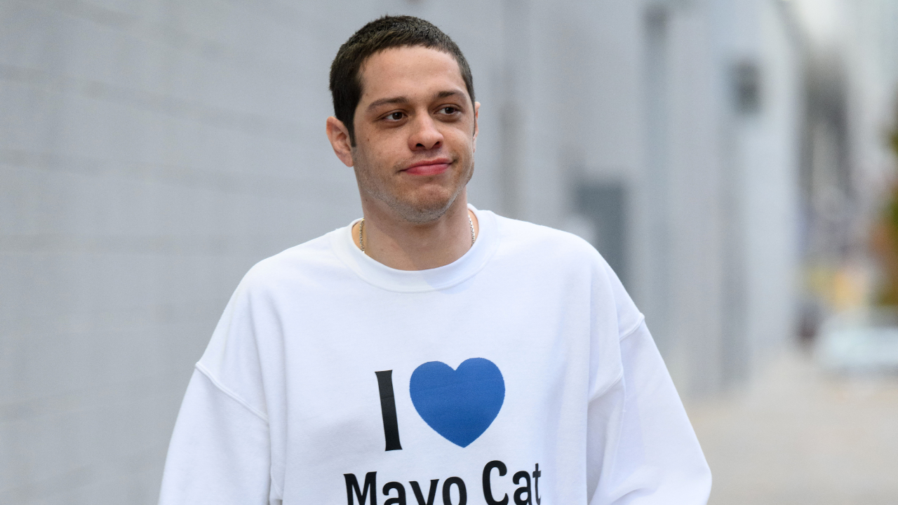 Pete Davidson wearing a white sweatshirt with "I [heart] Mayo Cat" printed on it, walking outside.