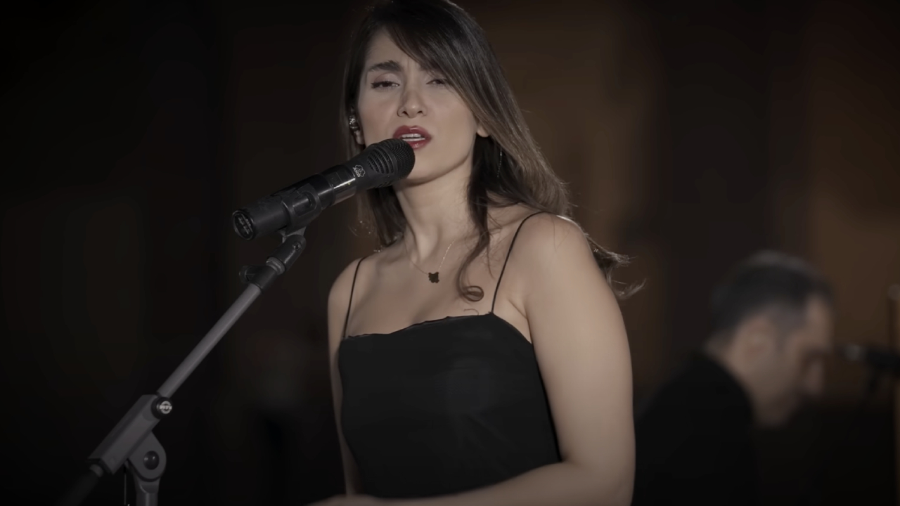Parastoo Ahmadi in a black dress sings into a microphone, with a musician blurred in the background.