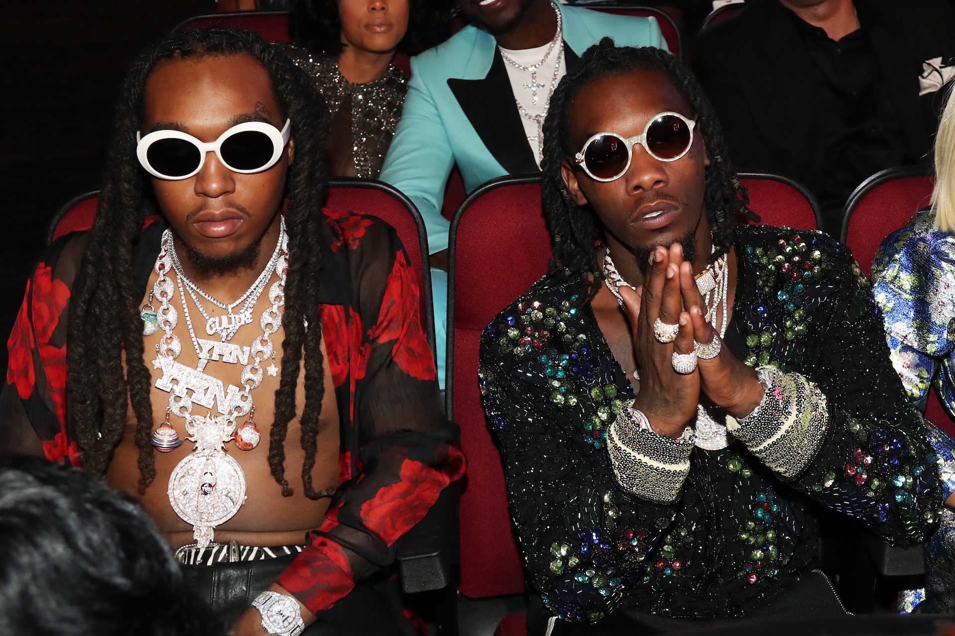 Takeoff and Offset from Migos sitting in an audience, wearing stylish outfits and sunglasses, adorned with jewelry.