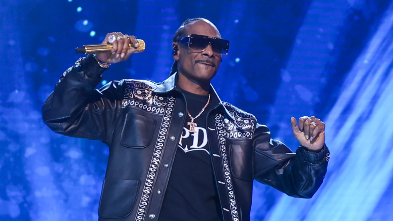 Snoop Dogg performing on stage, wearing sunglasses, a black leather jacket, and holding a gold microphone.