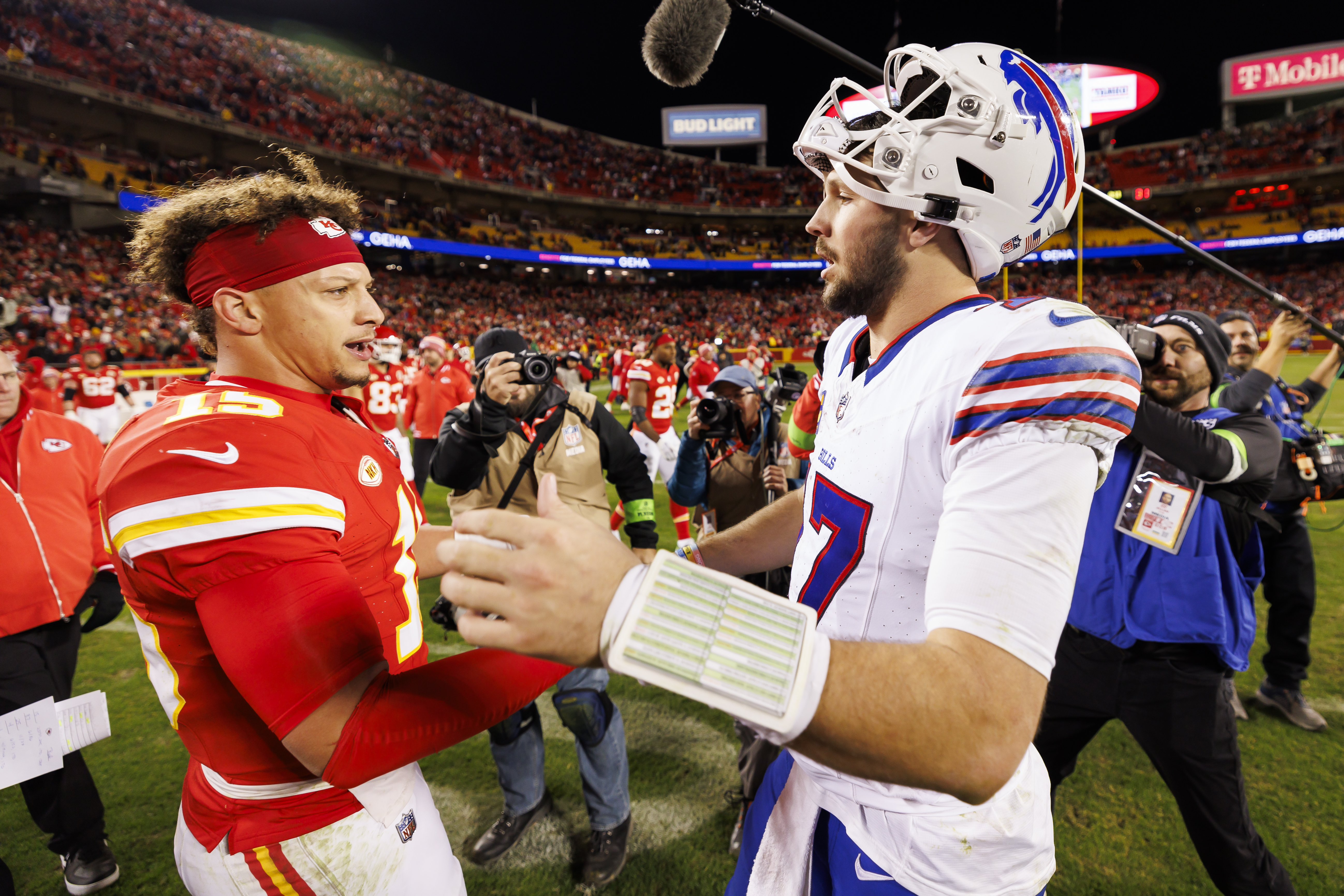 Patrick Mahomes and Josh Allen shaking hands on a football field, surrounded by photographers and fans in a stadium.