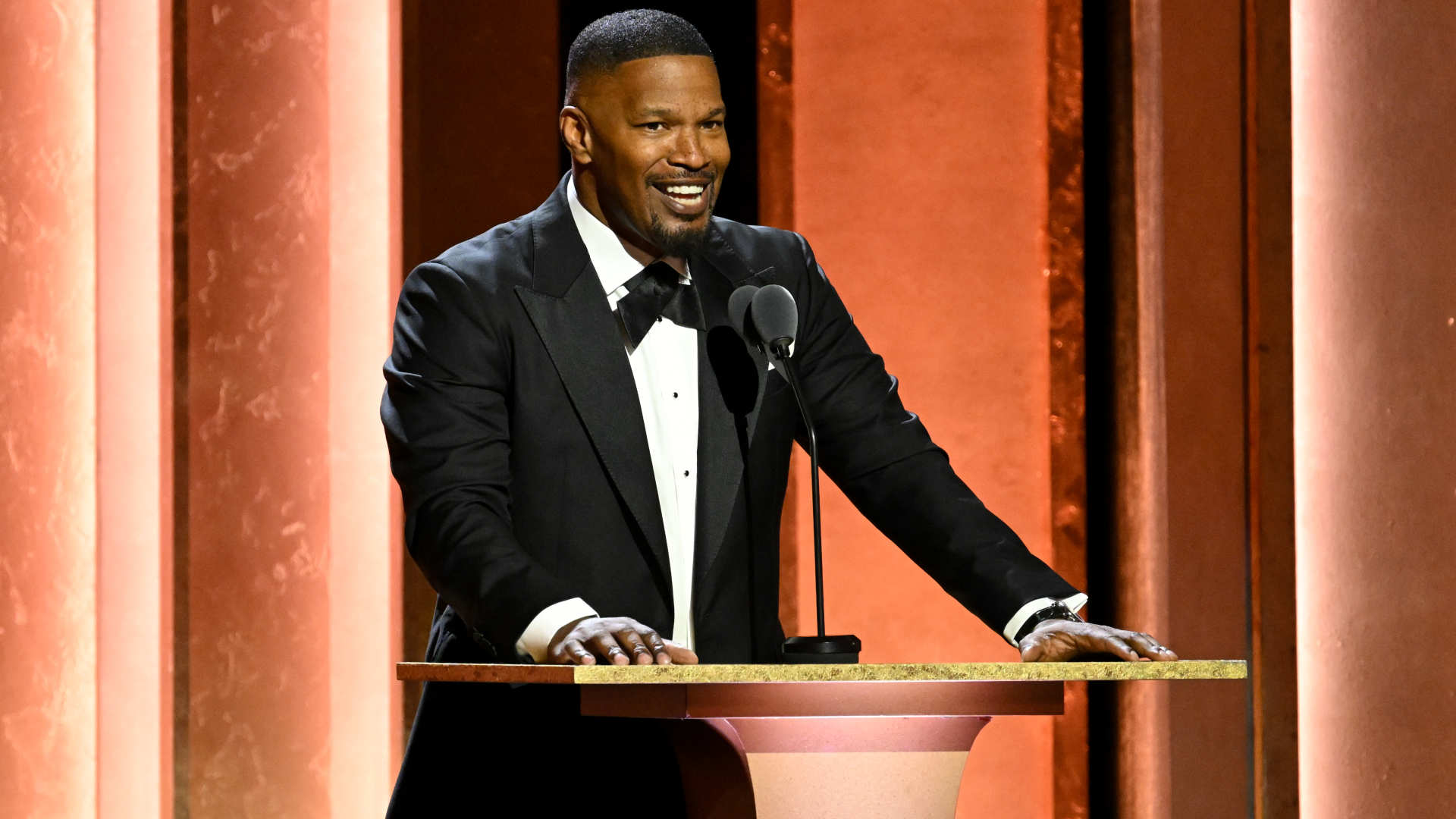 Jamie Foxx in a tuxedo, speaking at a podium with a warm, orange-lit background.