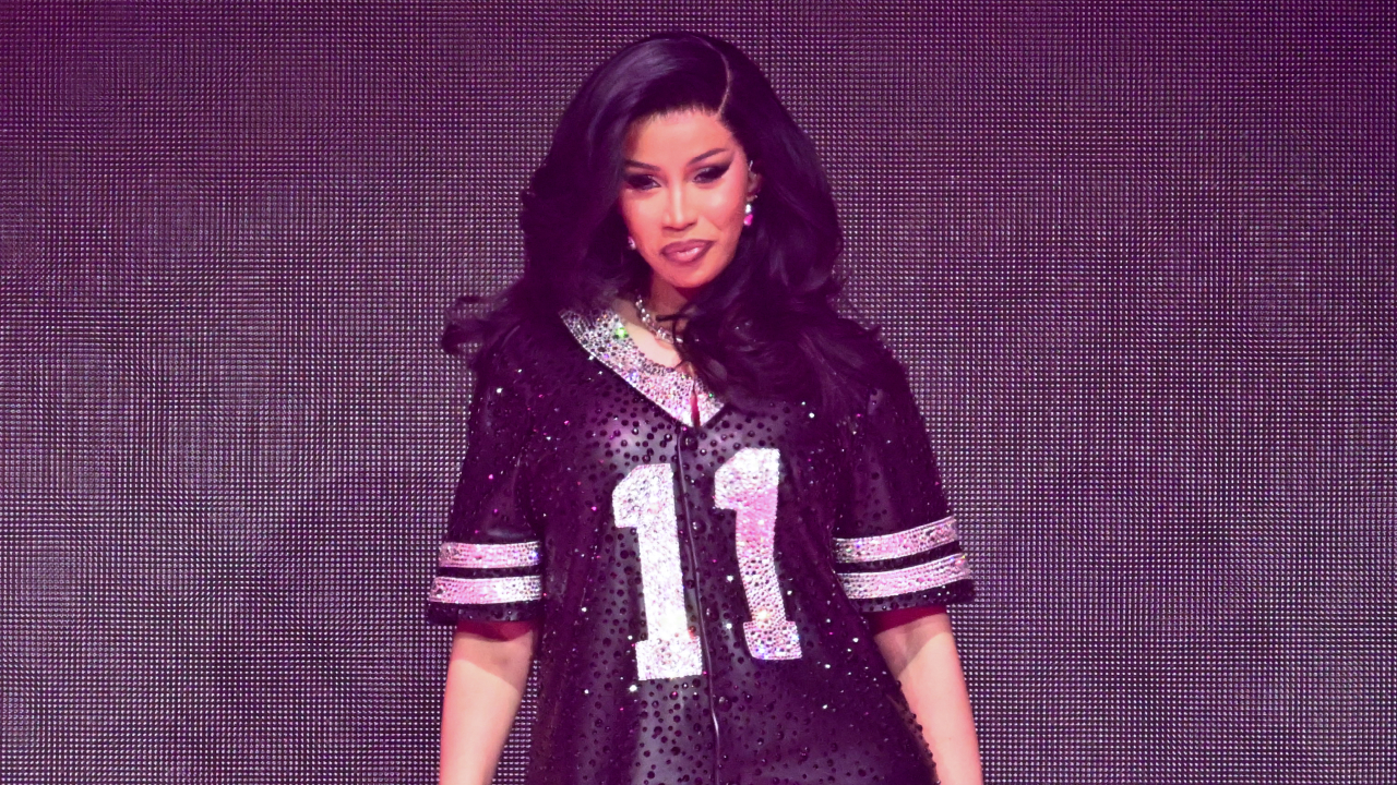 Cardi B performs on stage in a sparkly black jersey with the number 11, against a vibrant backdrop.