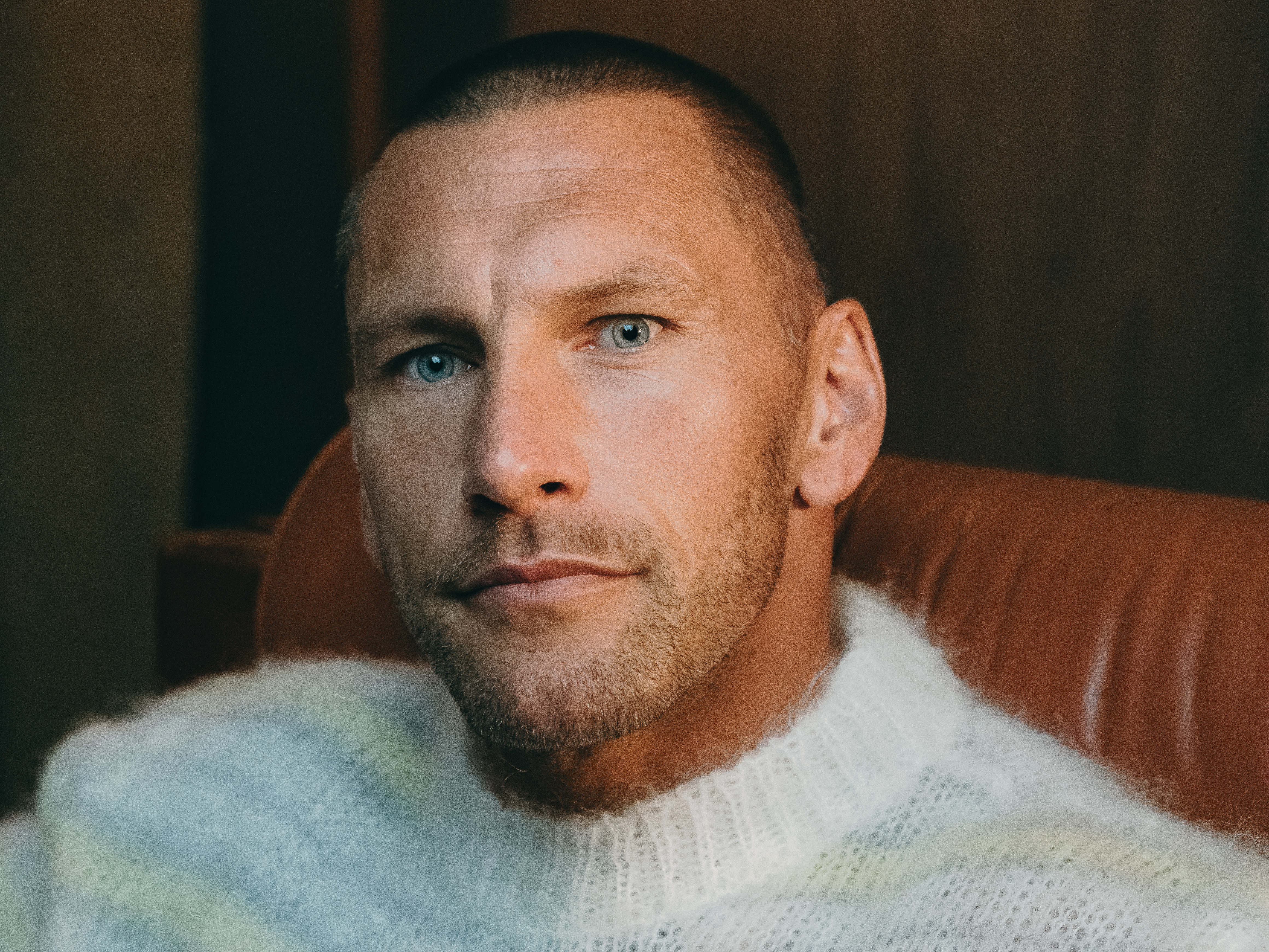 A man with short hair and a beard, wearing a fuzzy sweater, sits on a leather chair in a warmly lit room.
