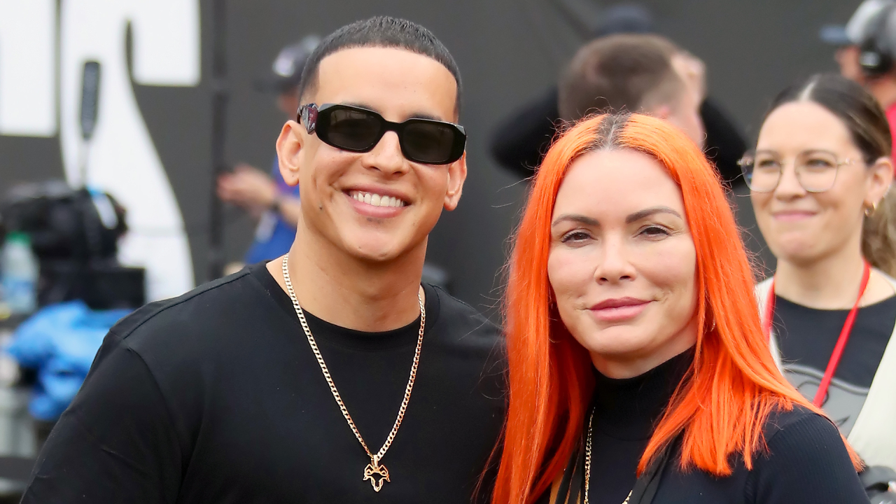 Daddy Yankee, wearing sunglasses and a black shirt, stands next to wife Mireddys González with bright orange hair. They are smiling at an event.