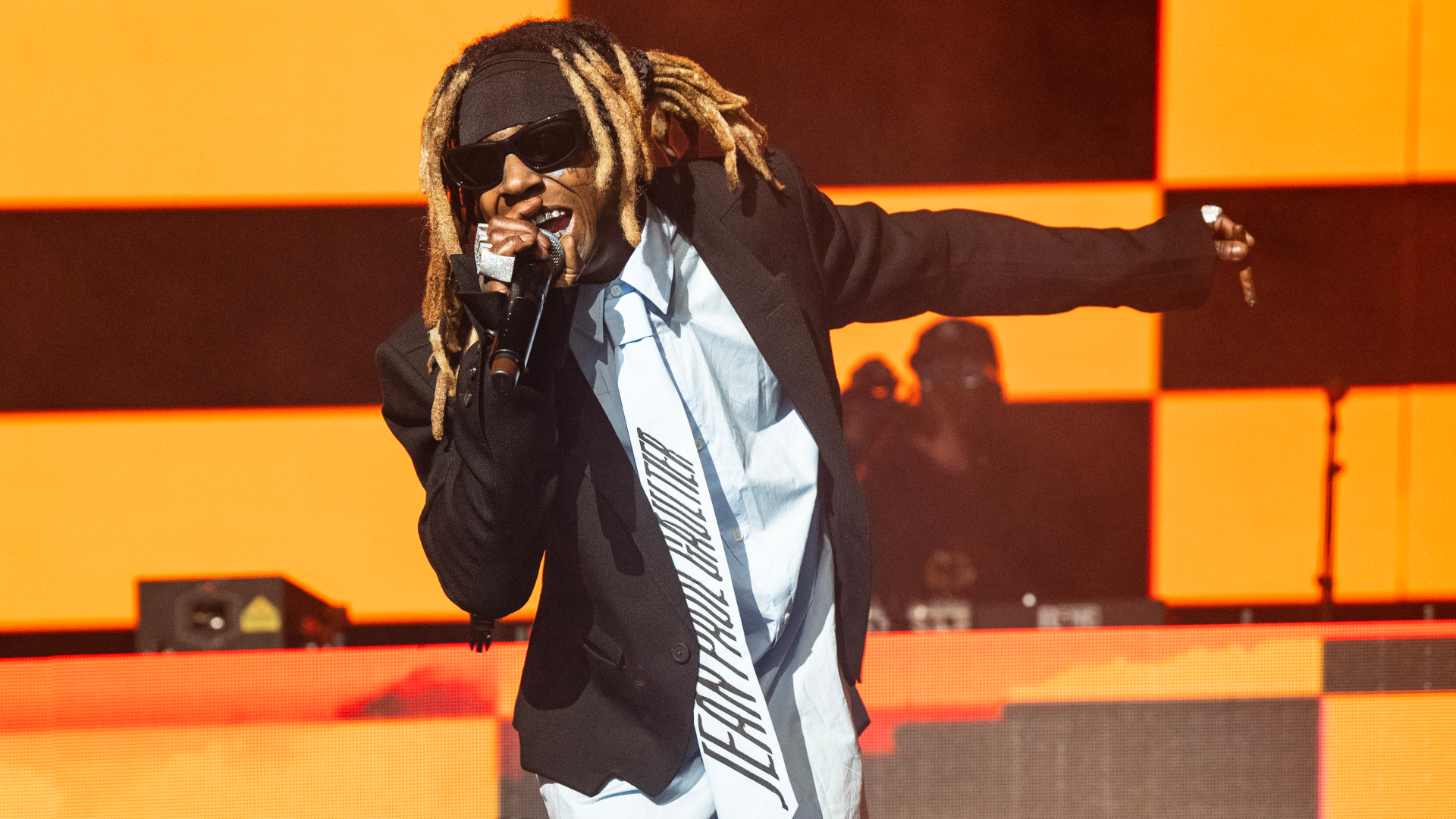 Weezy performing on stage with dreadlocks, sunglasses, and a black suit. The background features an orange and black checkered pattern.