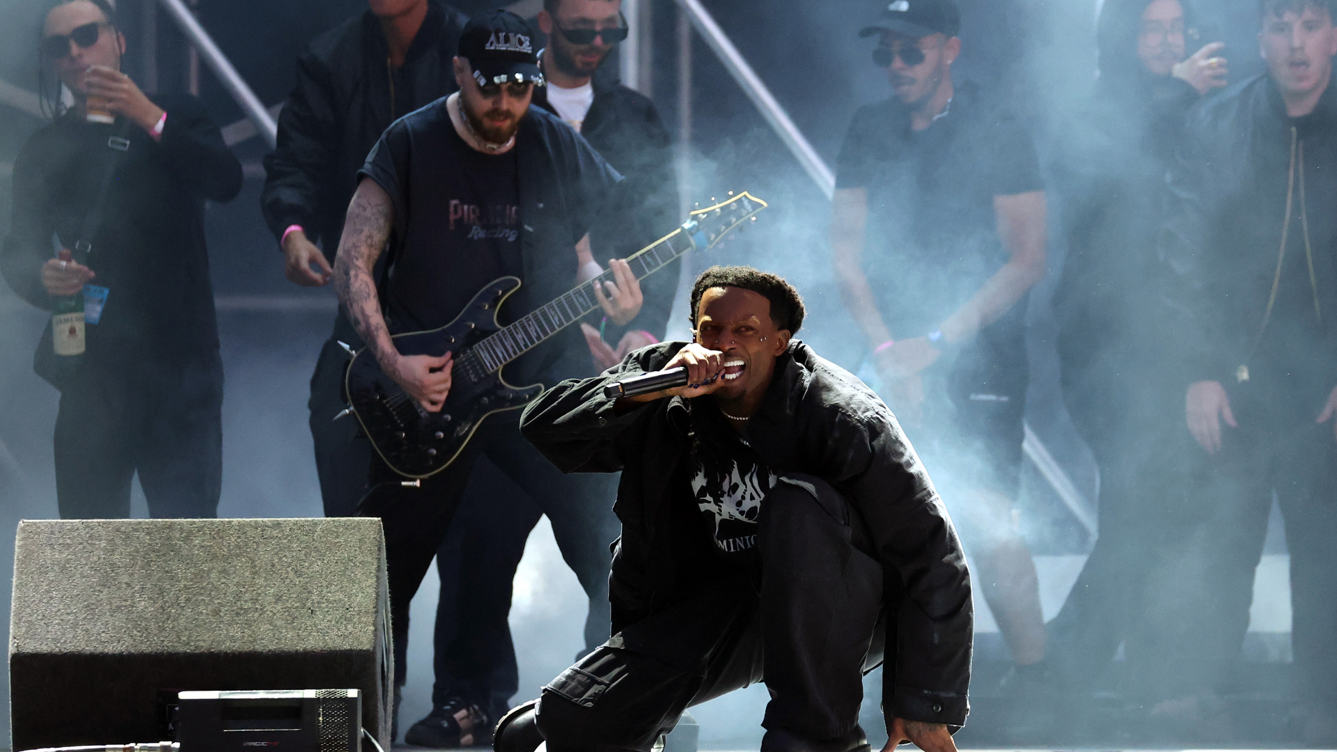 A dynamic stage performance with a singer crouching and holding a microphone, surrounded by musicians and a guitarist. Smoke fills the stage.