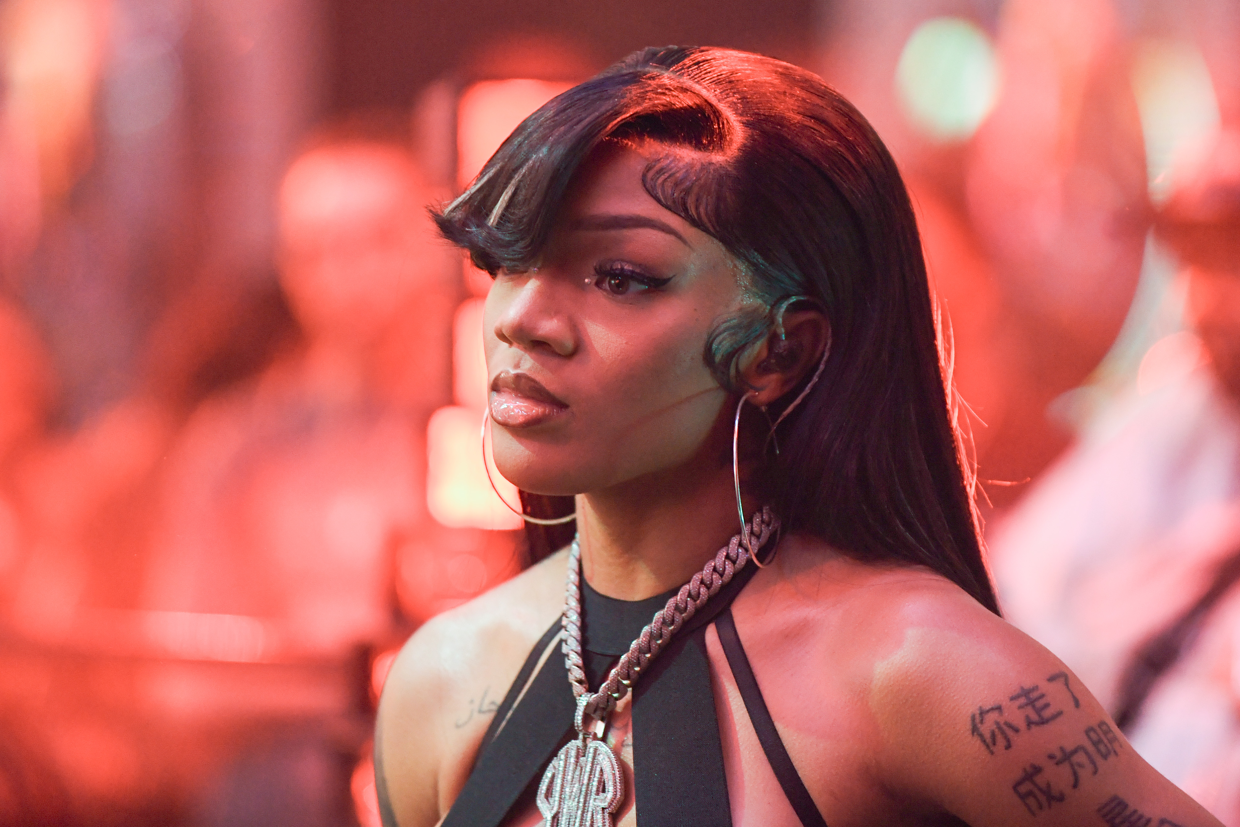 GloRilla with long hair and tattoos stands in a red-lit setting, wearing a halter top and large hoop earrings.