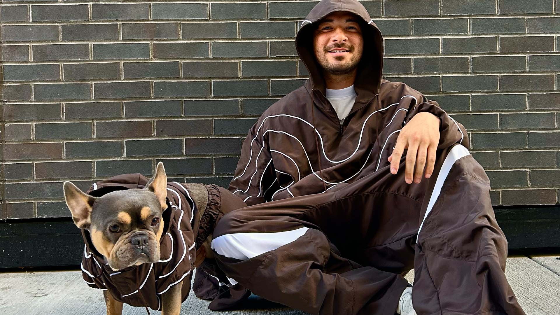 A person in a brown hooded tracksuit sits against a brick wall, smiling, with a French bulldog in a matching outfit beside them.