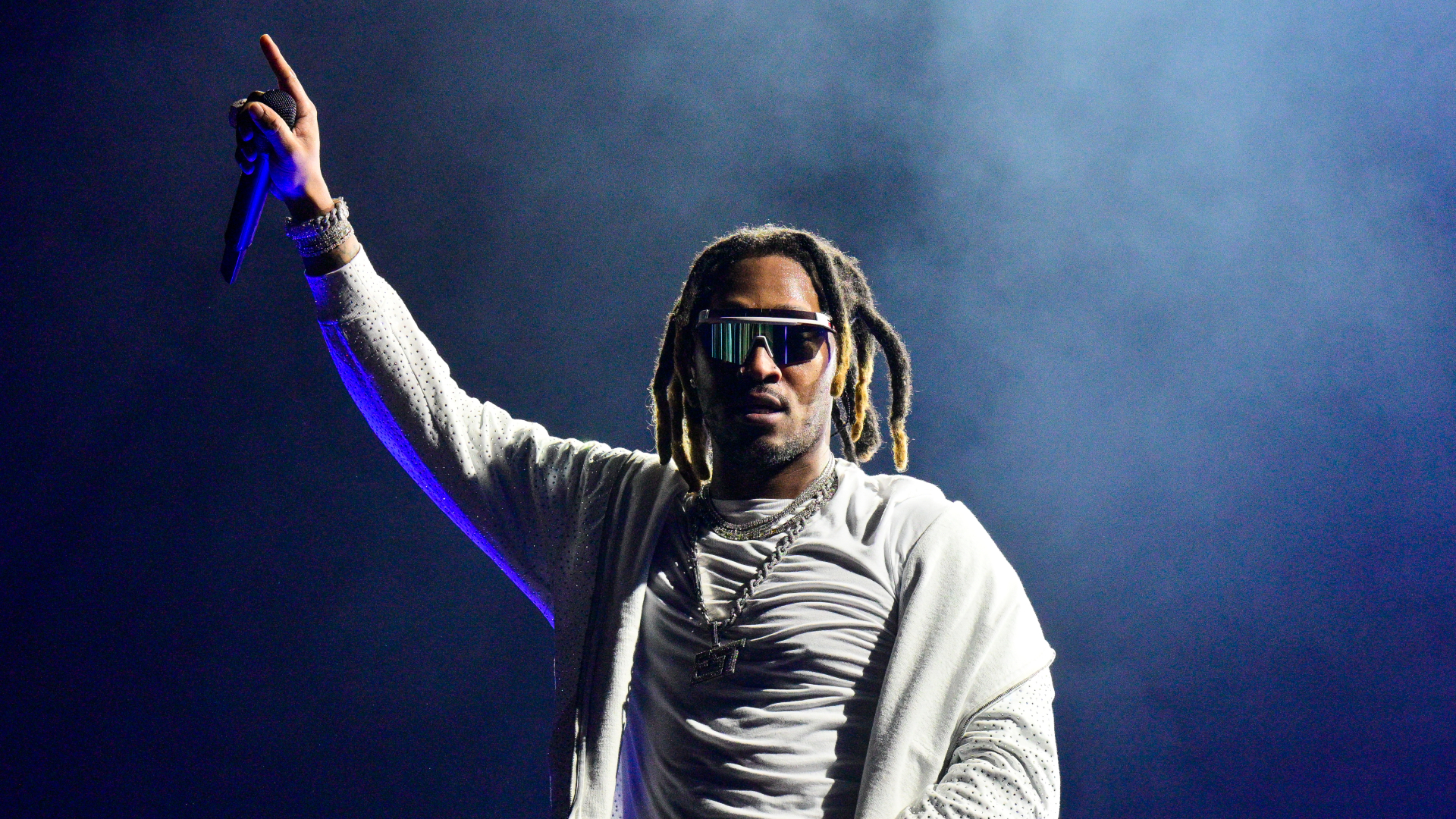 A performer on stage wearing sunglasses and a white outfit, holding a microphone with one hand raised, in front of a smoky background.
