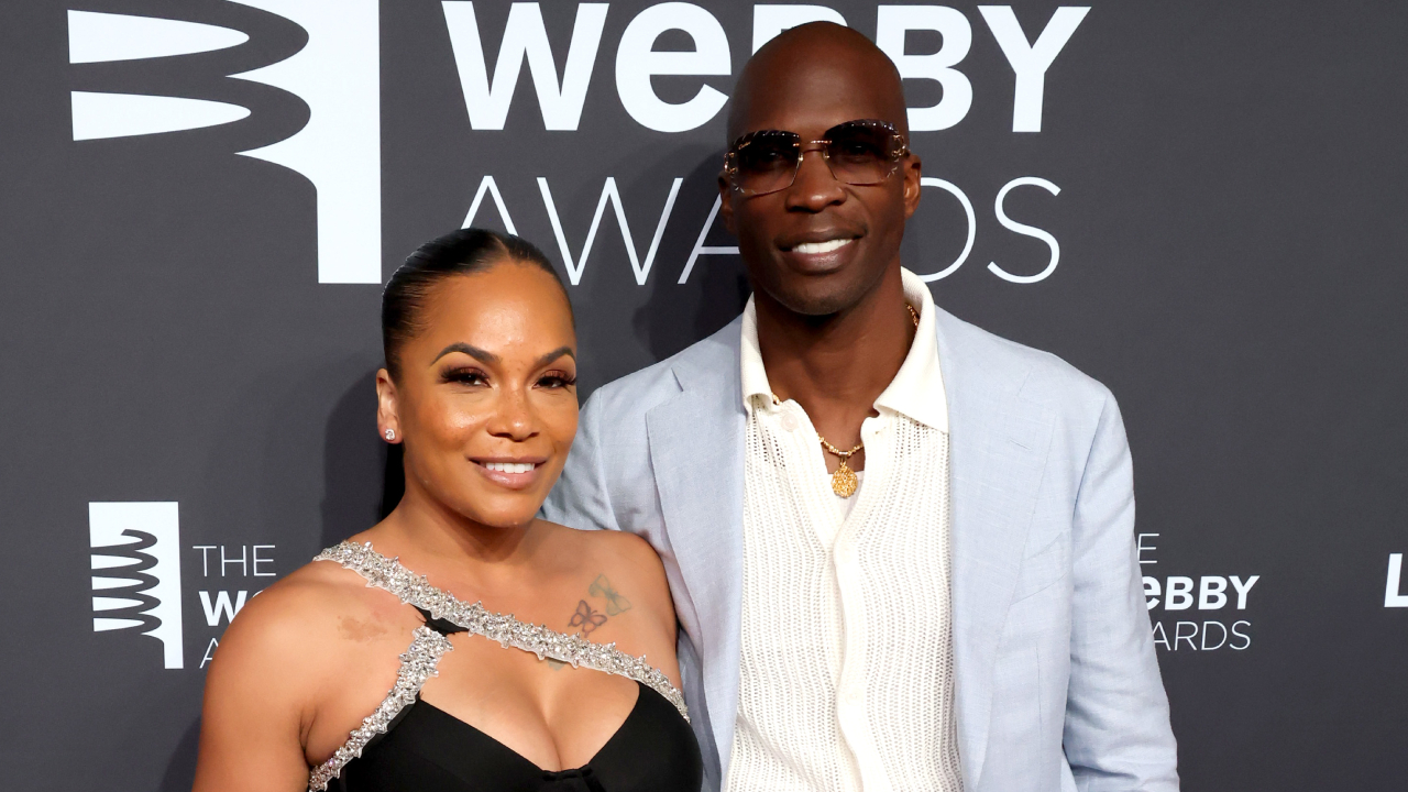Sharelle Rosado and Chad Ochocinco Johnson pose together. Rosado wears a black dress with silver accents, and Johnson wears a light blue suit and sunglasses.