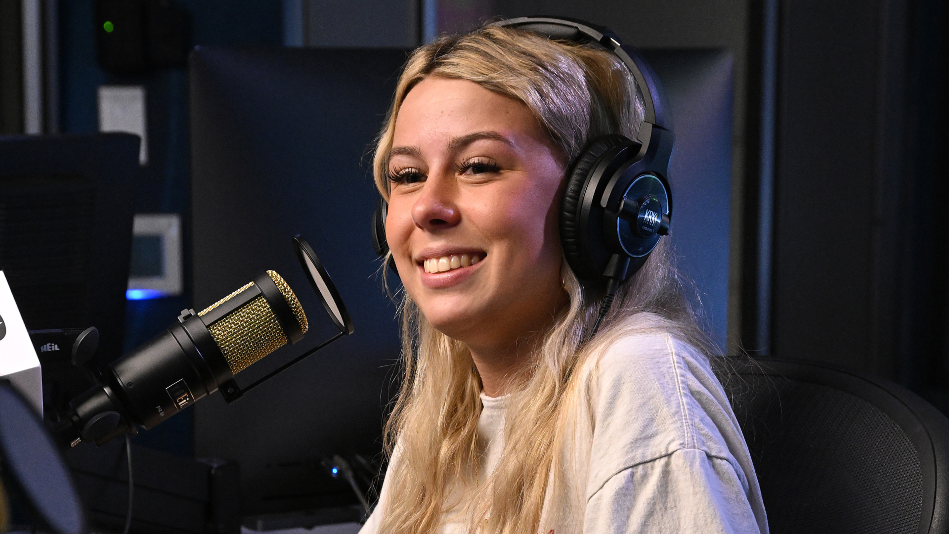 A woman with blonde hair and headphones is smiling while seated at a microphone in a recording studio.
