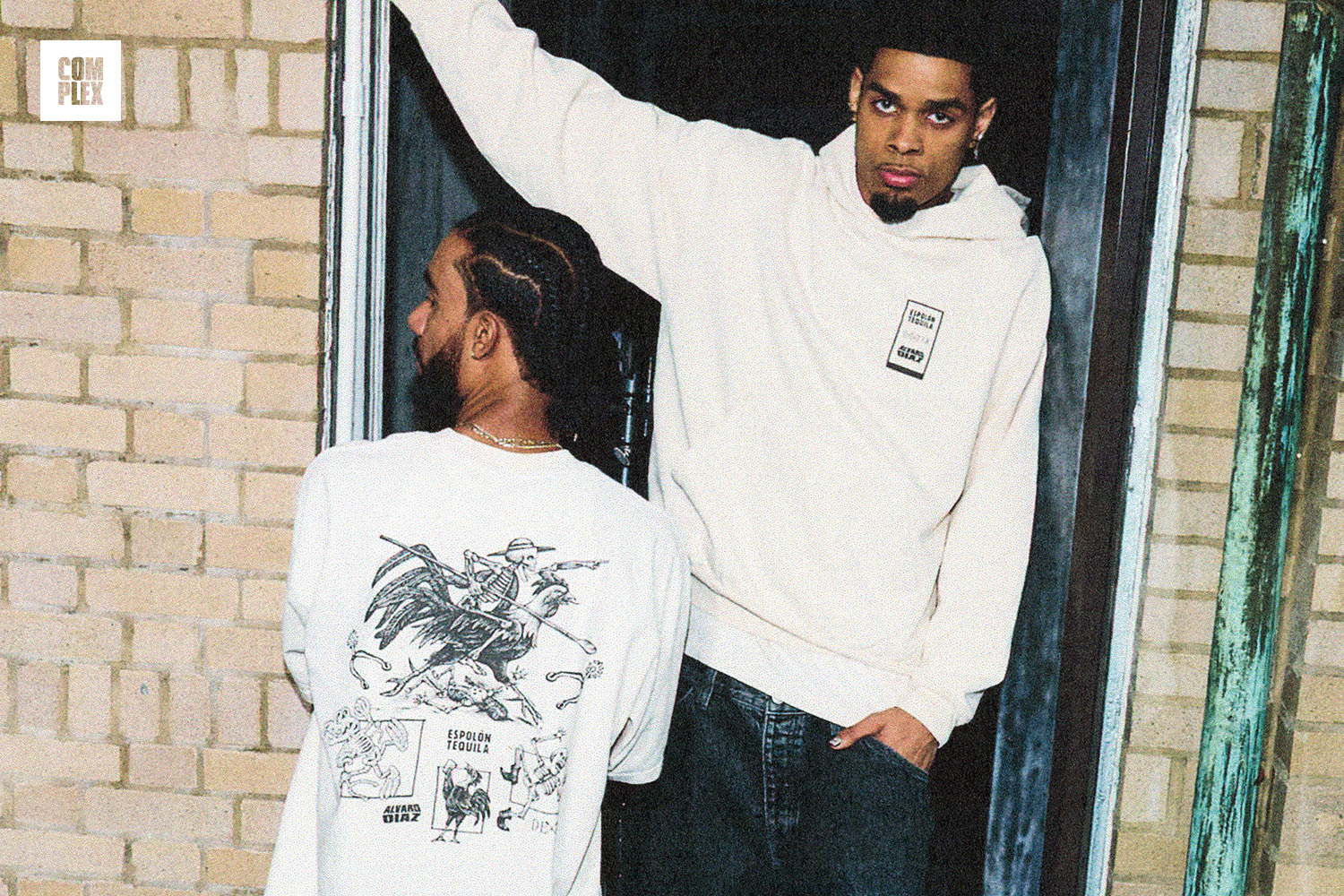 Two men in casual clothing stand by a brick wall. One wears a printed shirt, the other a hoodie. Complex logo is visible.