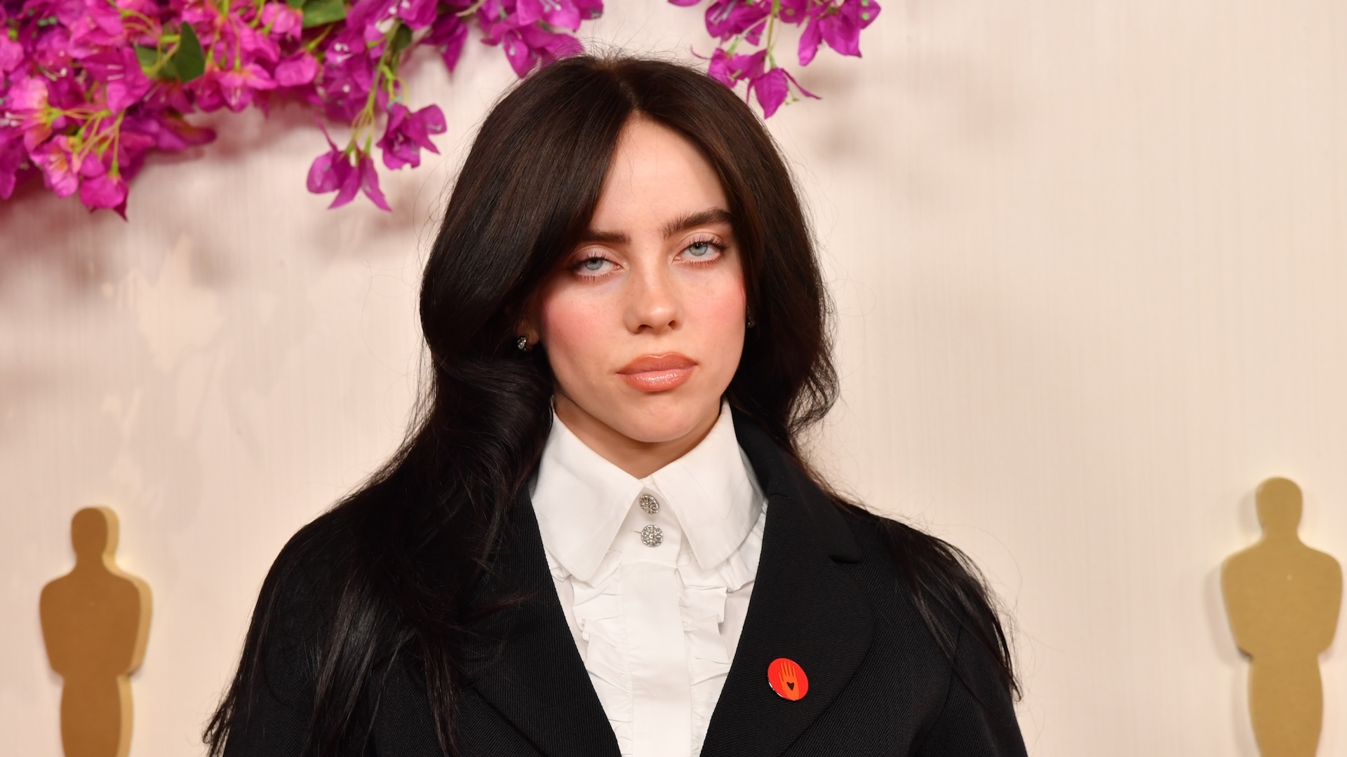 Billie Eilish at an event, wearing a black suit with a white shirt, surrounded by Oscar statues and purple flowers.