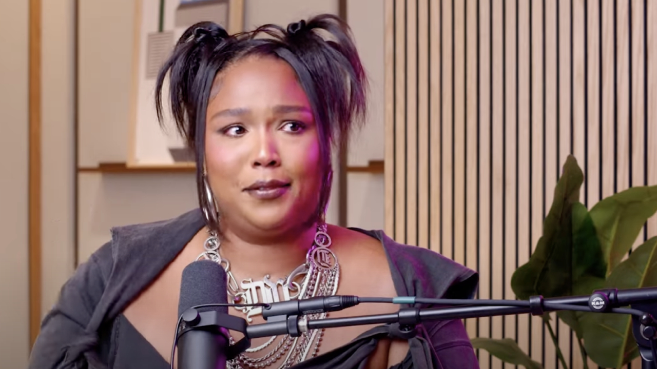 Lizzo speaking into a microphone, wearing a layered necklace and a dark outfit, with her hair styled in two buns.