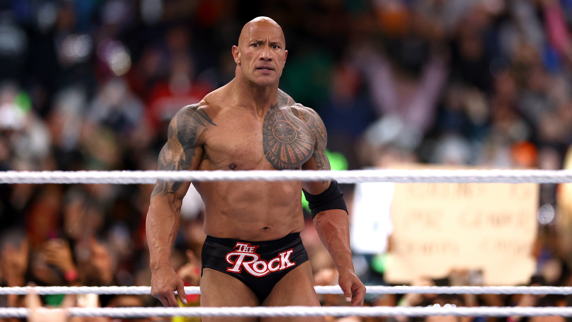 Dwayne "The Rock" Johnson in a wrestling ring, wearing black trunks with "The Rock" written on them, surrounded by a cheering crowd.