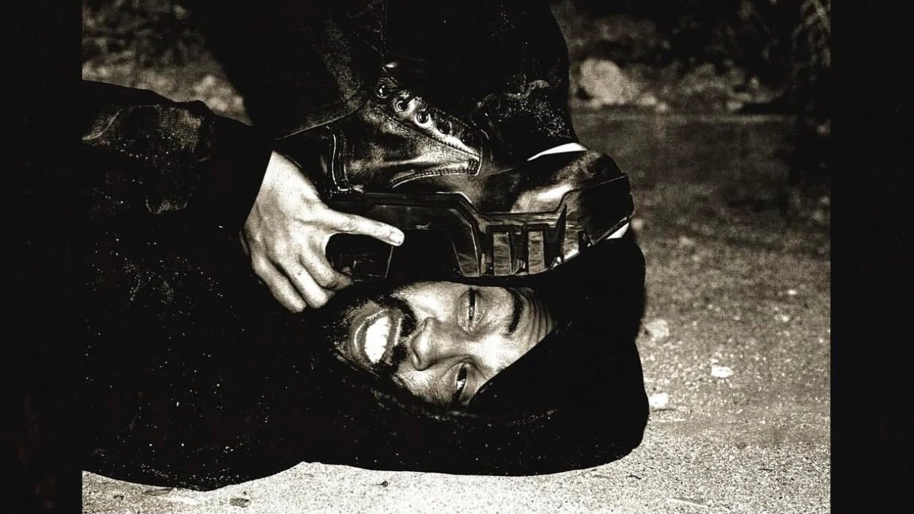 A man lying on the ground with a boot pressed against his face, smiling. The image is in black and white.