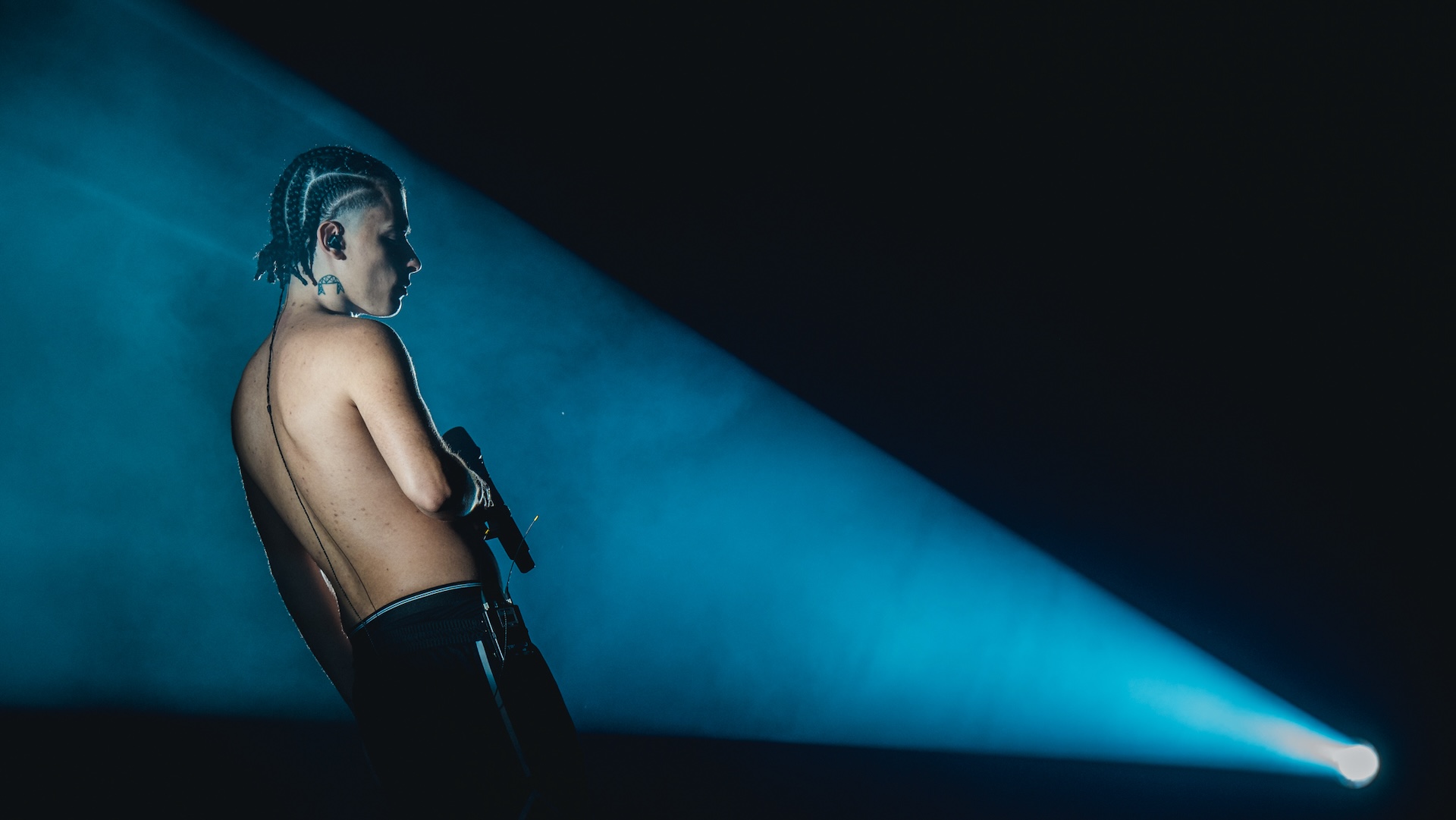 Trueno shirtless performs with braided hair stands on stage, holding a microphone, illuminated by a blue spotlight.
