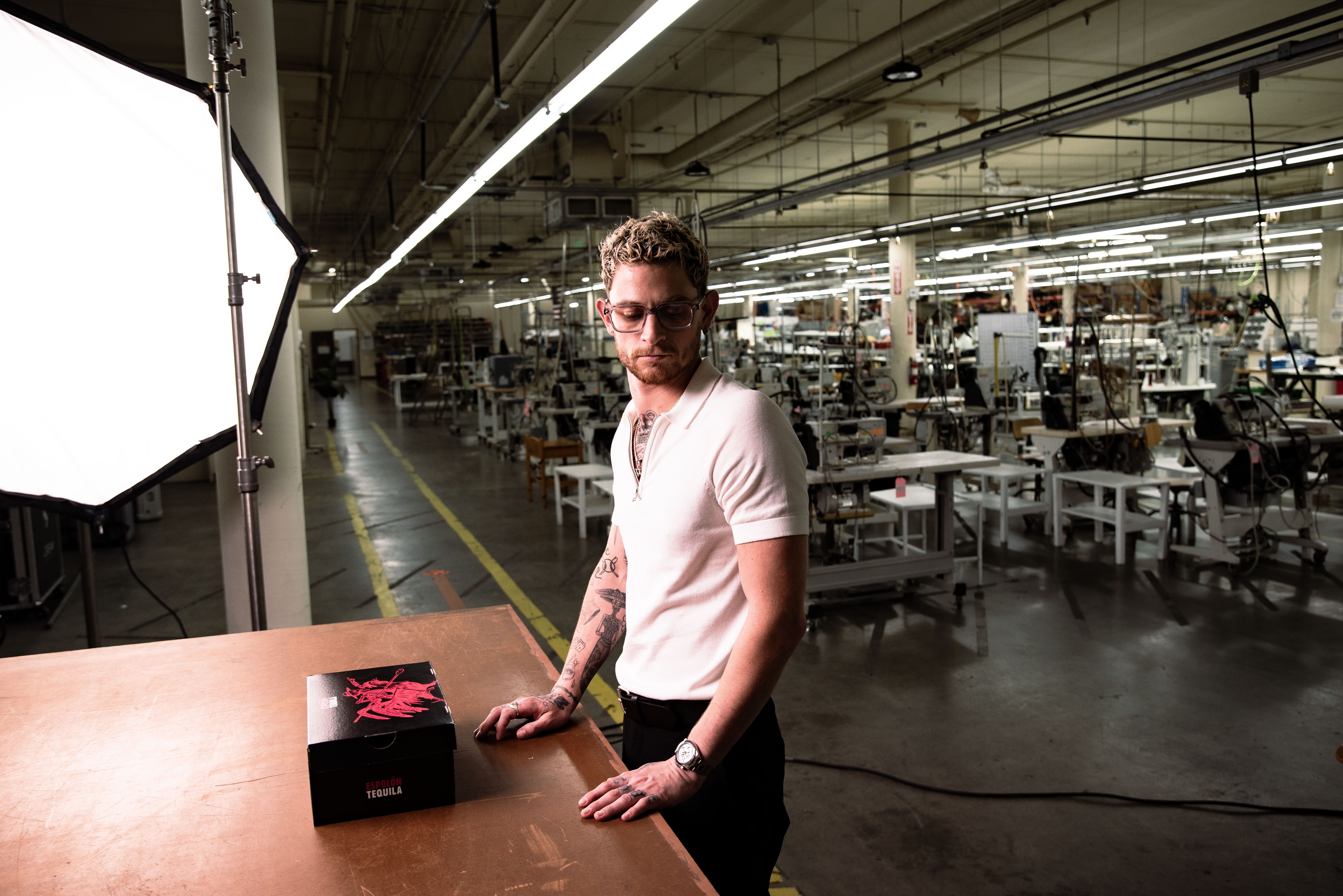 A man with glasses and tattoos stands in a factory, leaning on a table with a black box featuring a pink design.