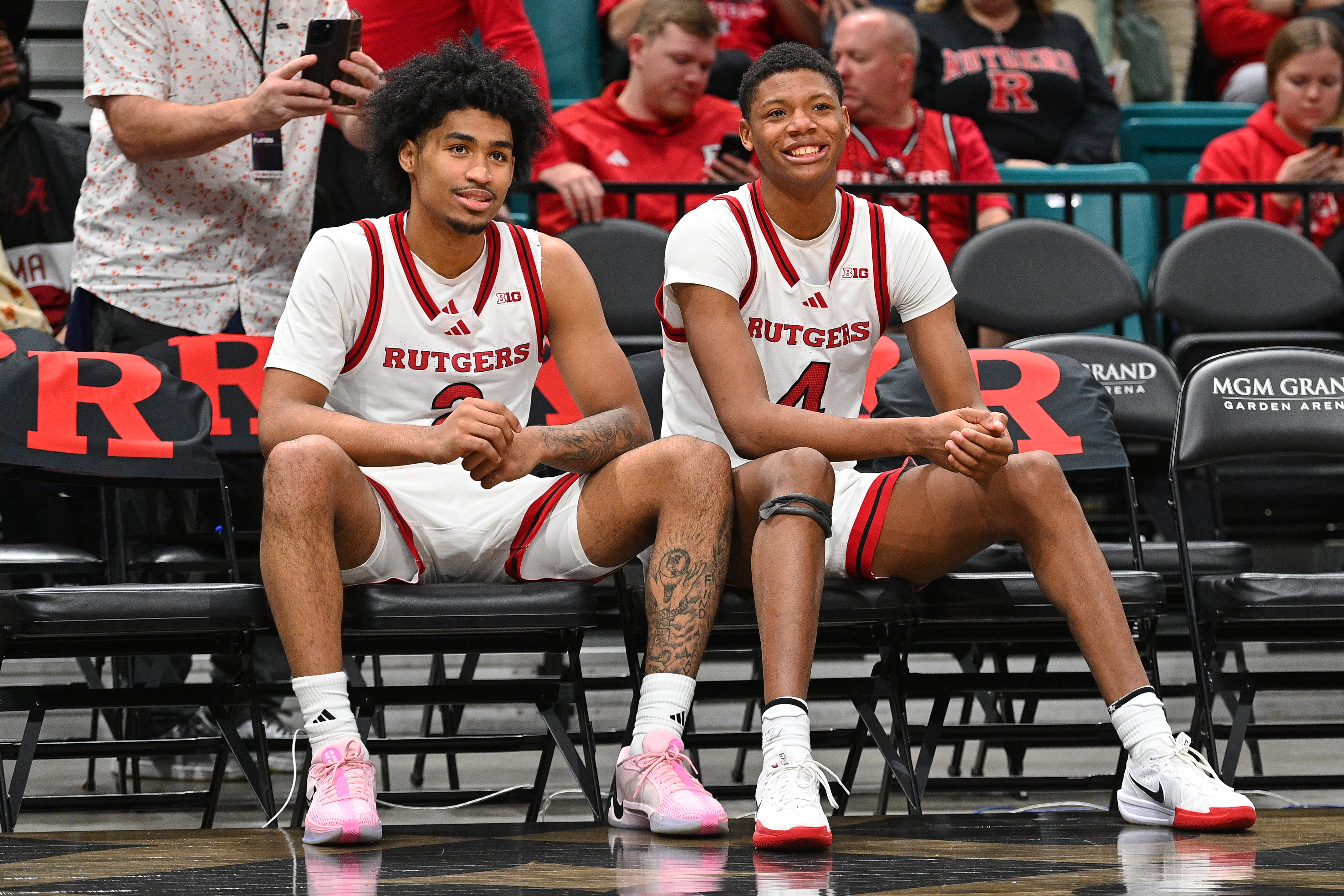 Rutgers Scarlet Knights guard Dylan Harper (2) and Rutgers Scarlet Knights guard Ace Bailey (4) sitting on the bench, smiling, wearing white and red uniforms. Spectators in the background.