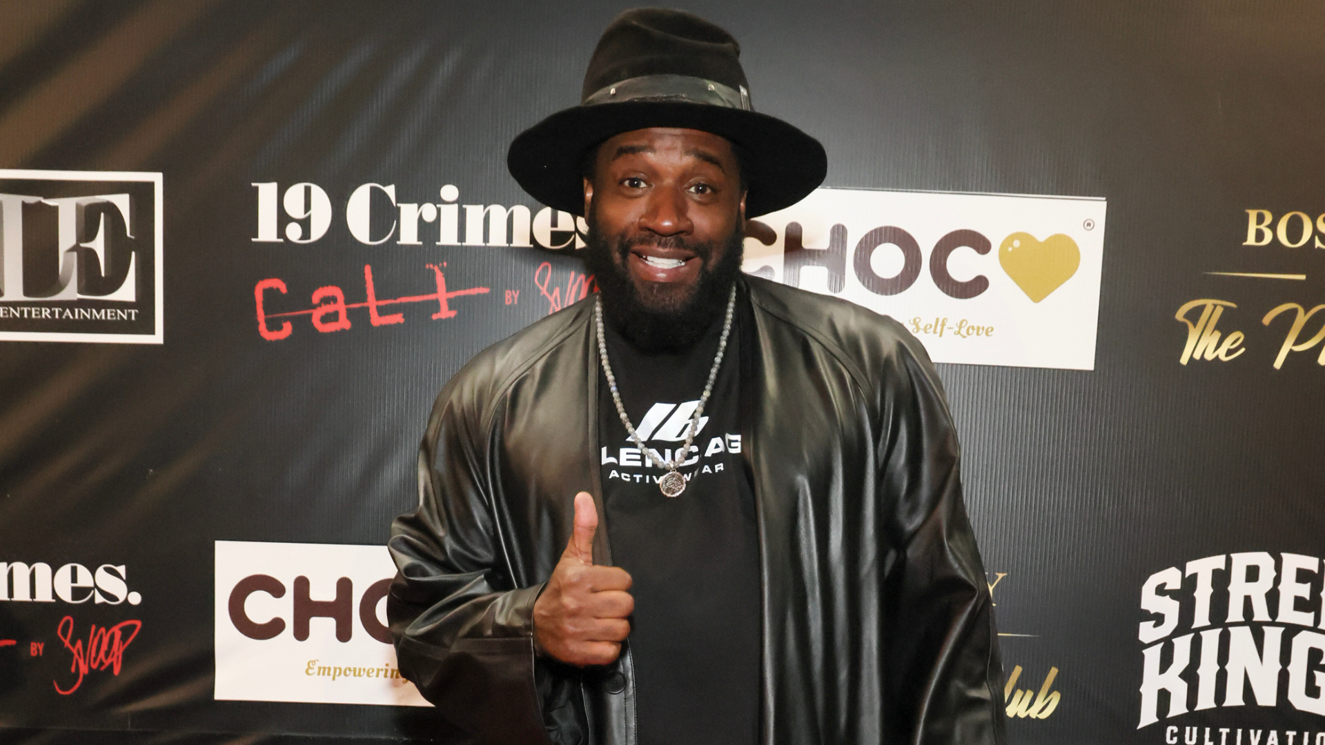 A man in a black hat and leather jacket gives a thumbs up at a promotional event backdrop.