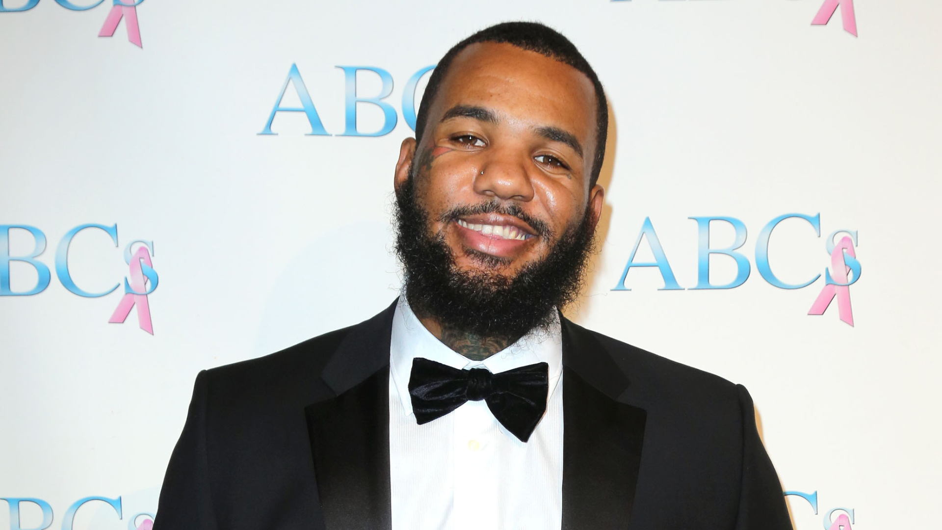 A man in a tuxedo with a bow tie smiles at an event, standing in front of a backdrop with "ABC's" and pink ribbons.