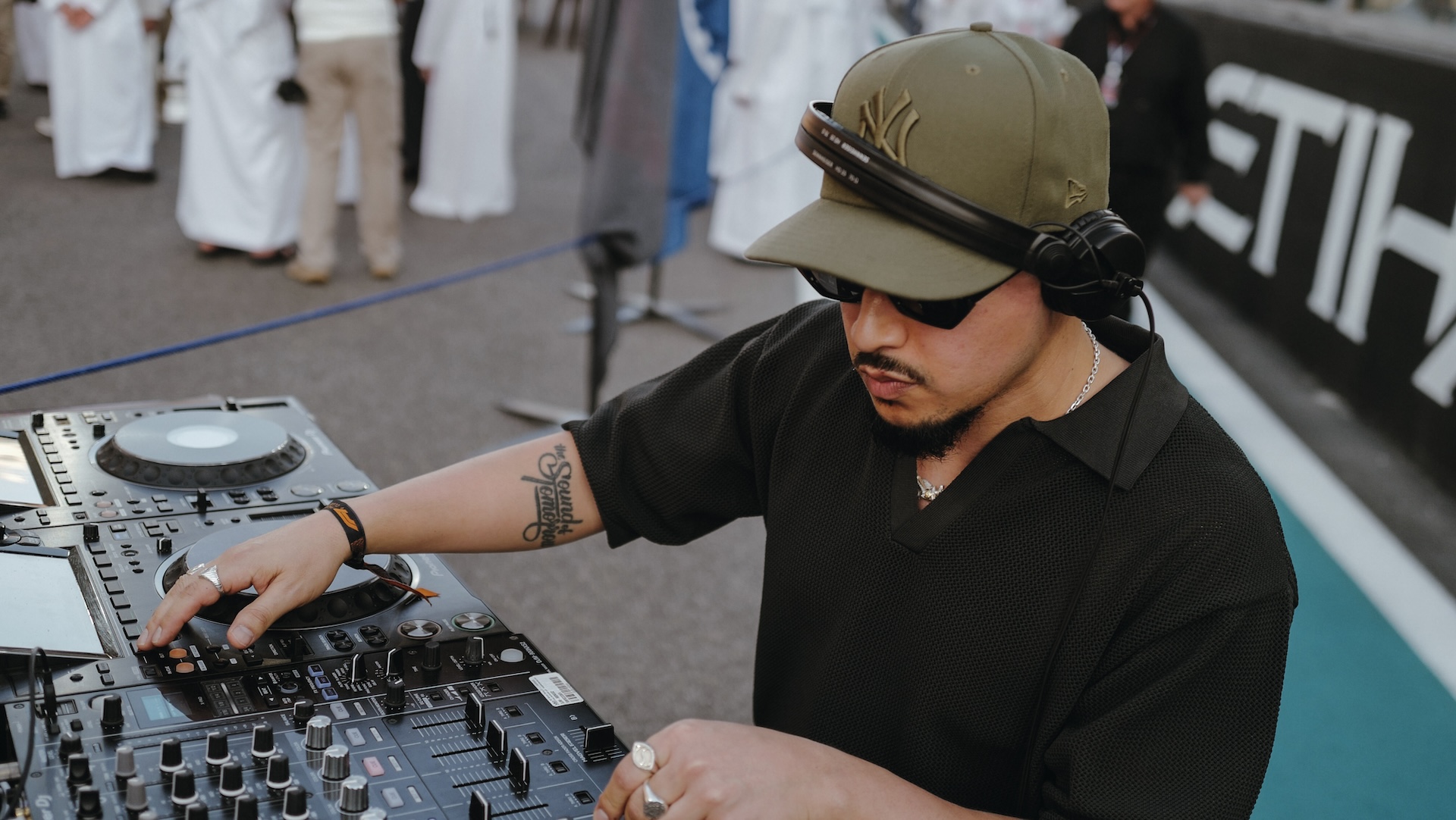 Joe Kay is wearing a cap and sunglasses is mixing music on a turntable at an outdoor event. People in white robes are in the background.